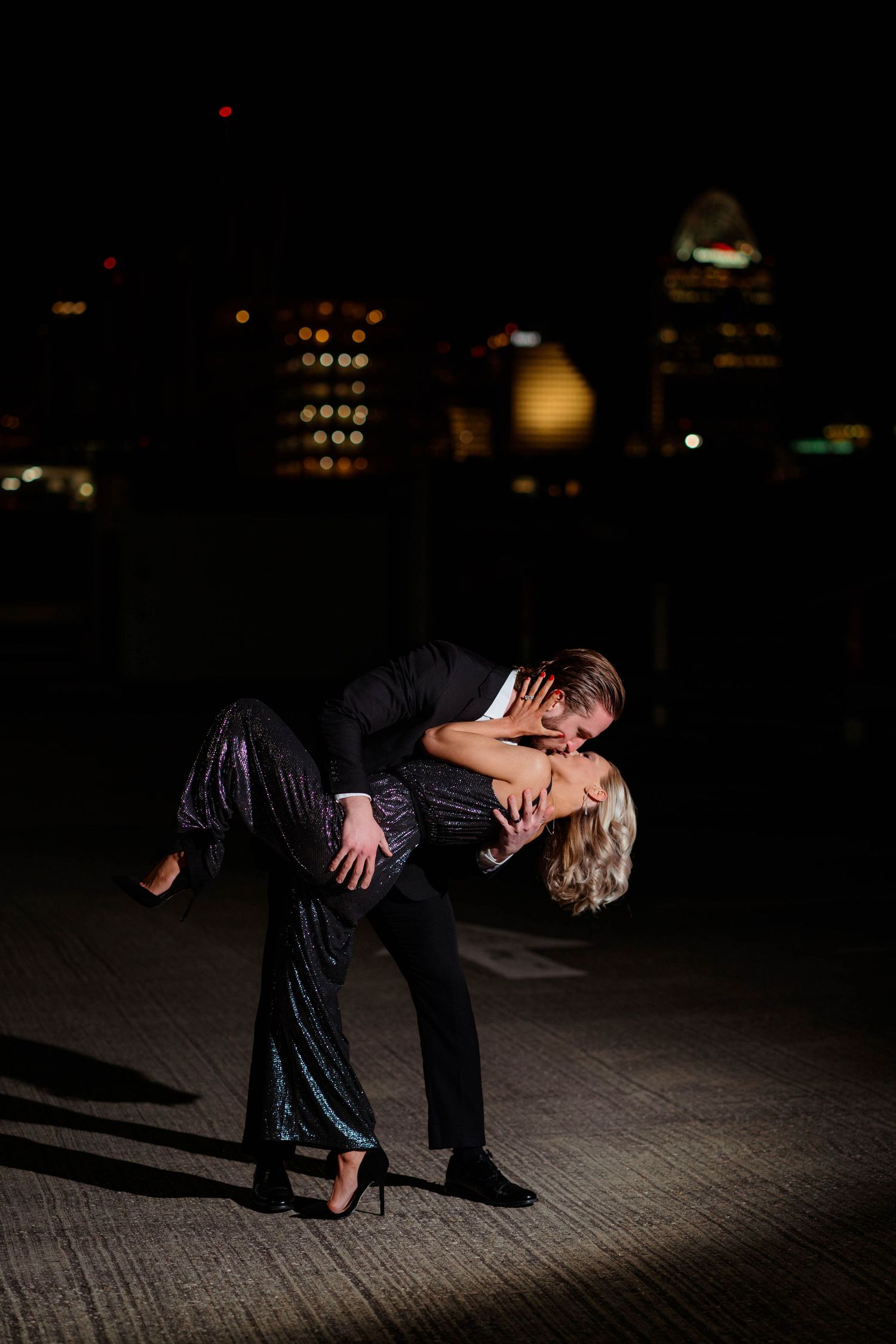 Couple performing a dramatic dip kiss at night with glowing city lights visible in the dark background.