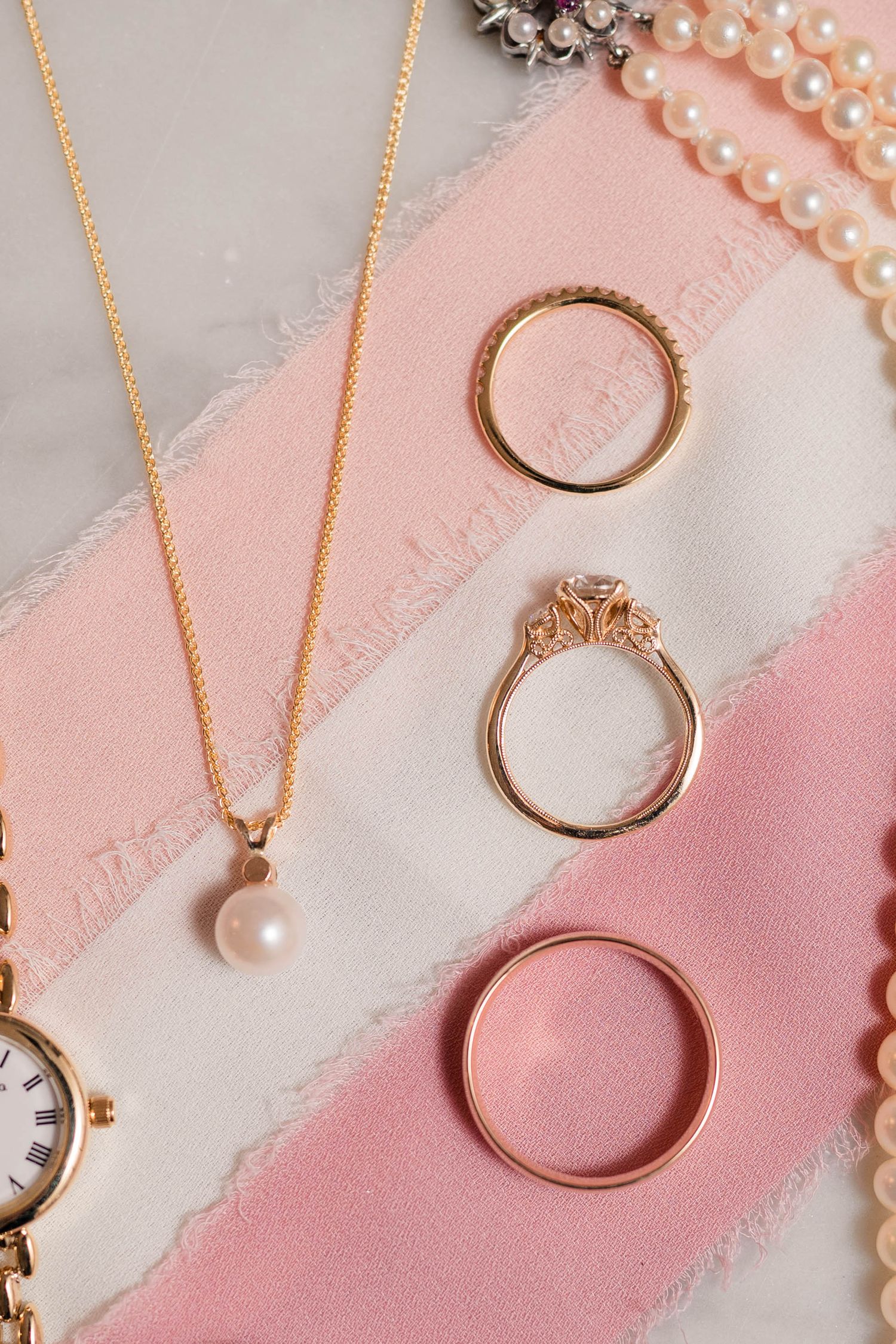 Gold wedding rings, diamond engagement ring, and pearl pendant necklace arranged on pink ribbon.