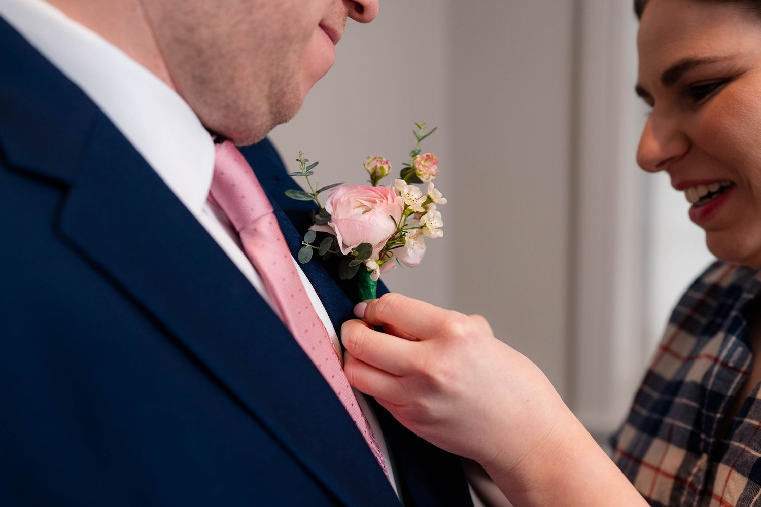 Close-up of a pink rose boutonniere being pinned to a navy wedding suit lapel.