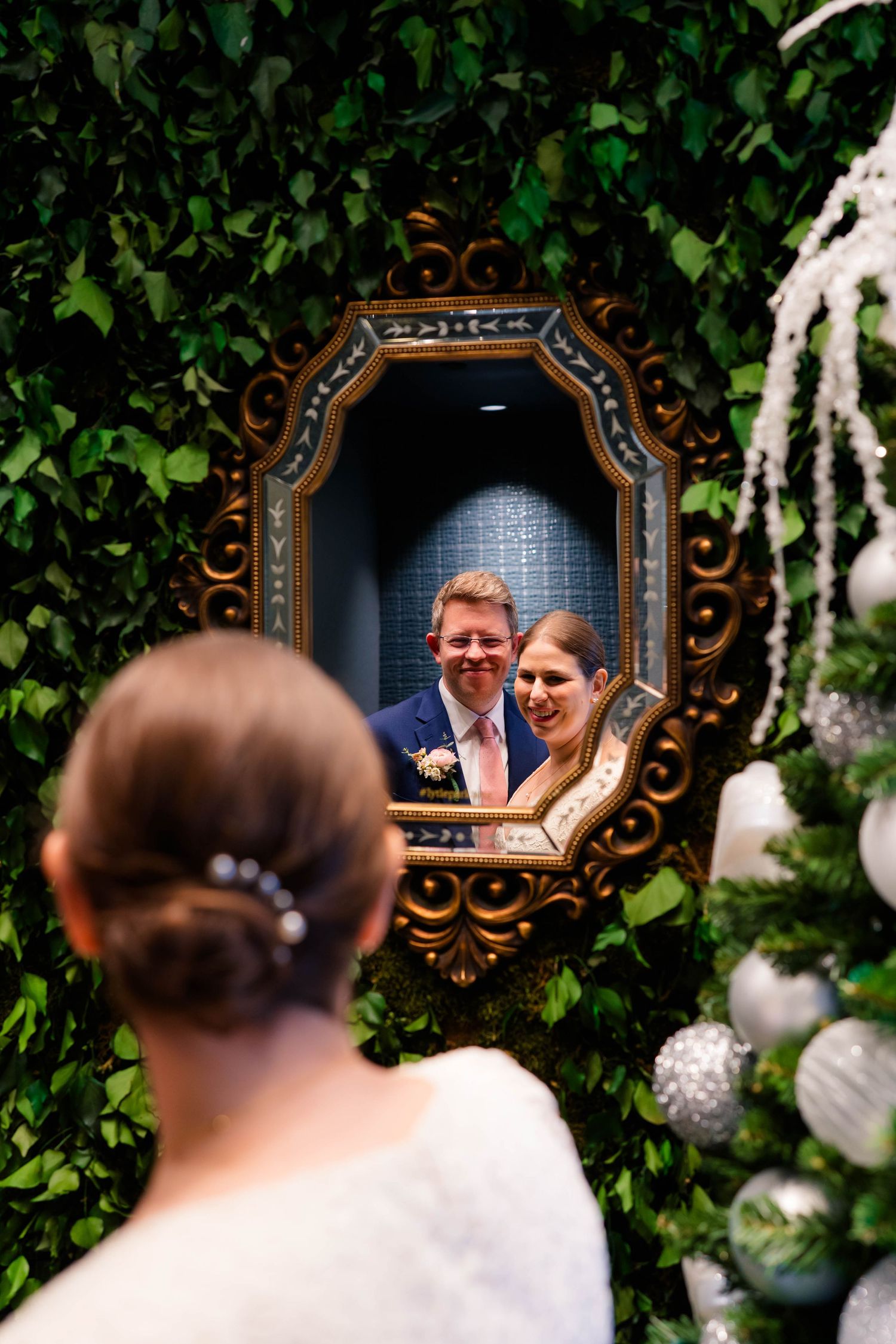 Wedding couple smiles together reflected in an ornate gold mirror surrounded by lush green ivy wall decor.