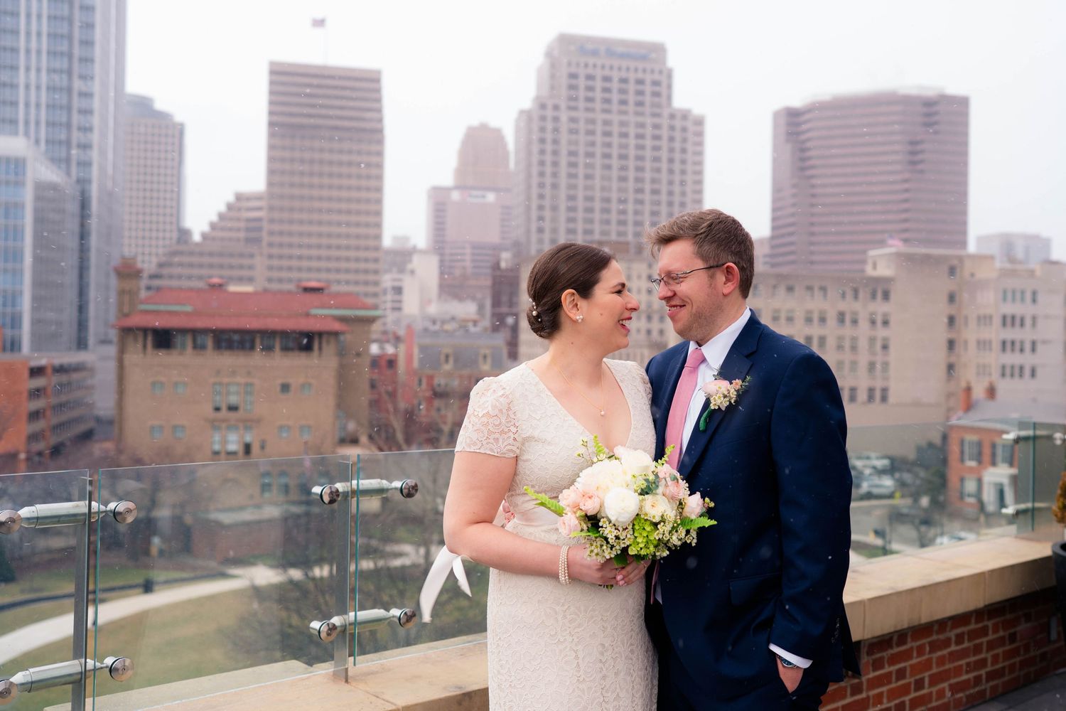 Bride and groom sharing a romantic glance on rooftop venue with misty urban skyline behind them.