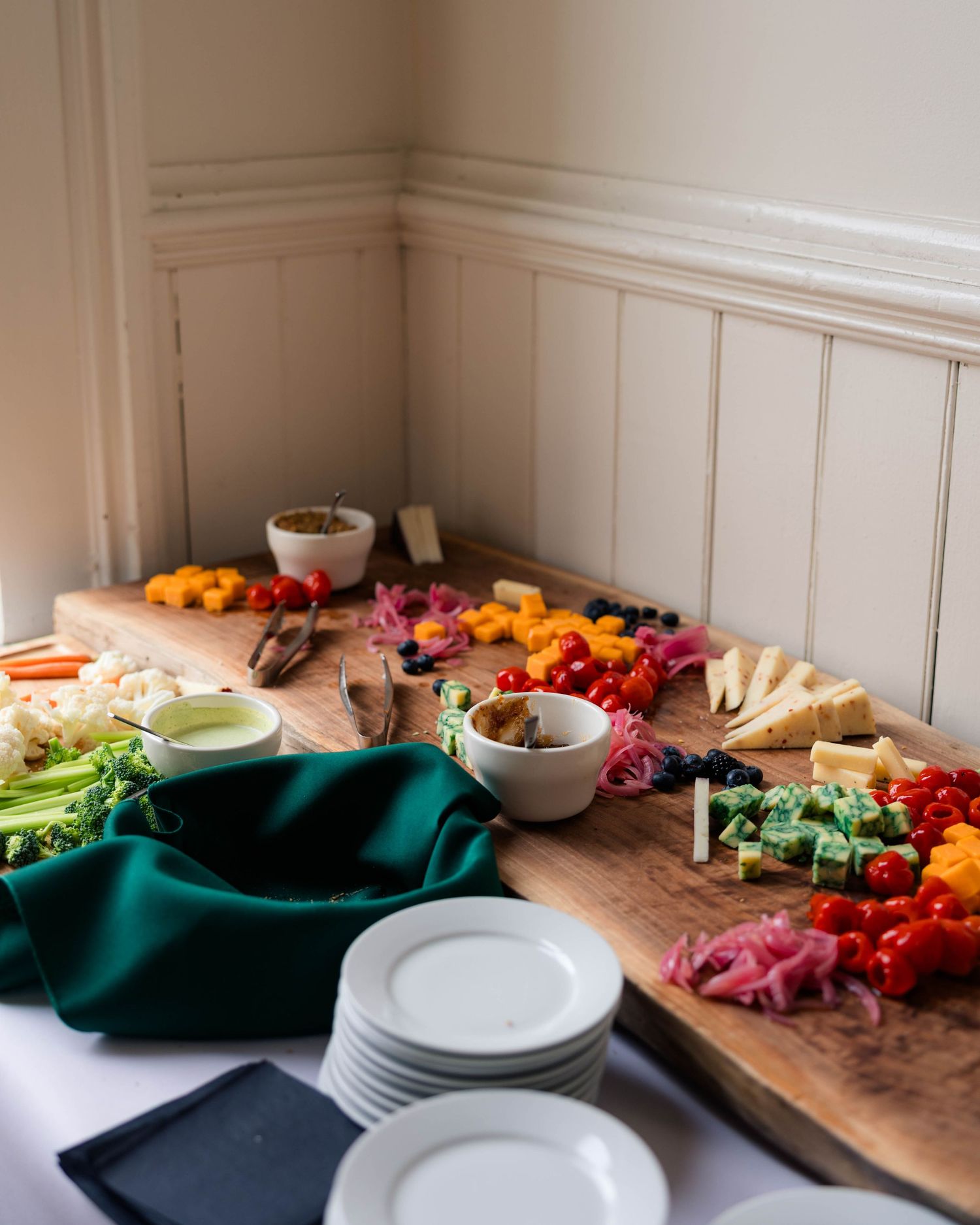 Rustic wooden charcuterie board with colorful cheeses, vegetables, and white plates at a wedding.