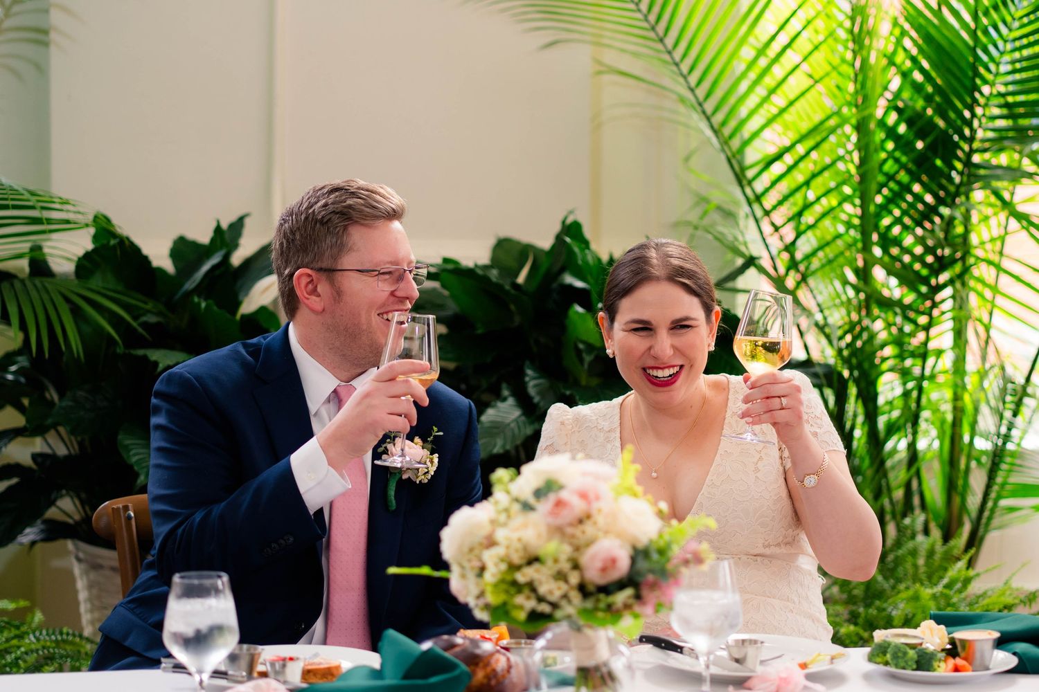 Bride and groom laughing and clinking champagne glasses at their wedding reception dinner table.