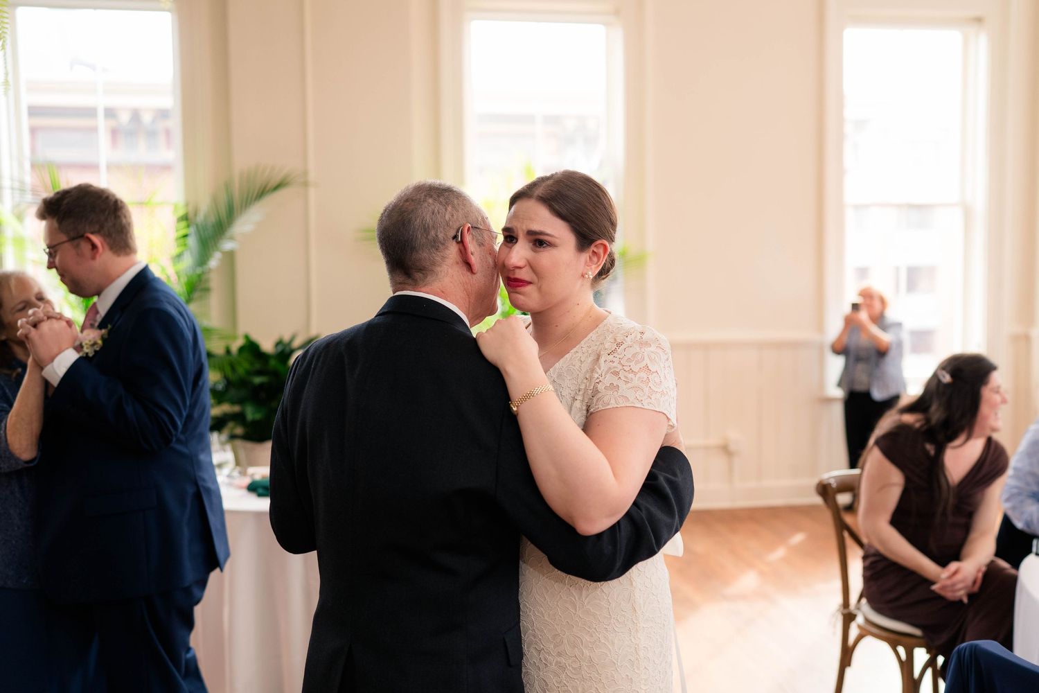 Emotional bride in lace dress wiping tears during father-daughter wedding dance.