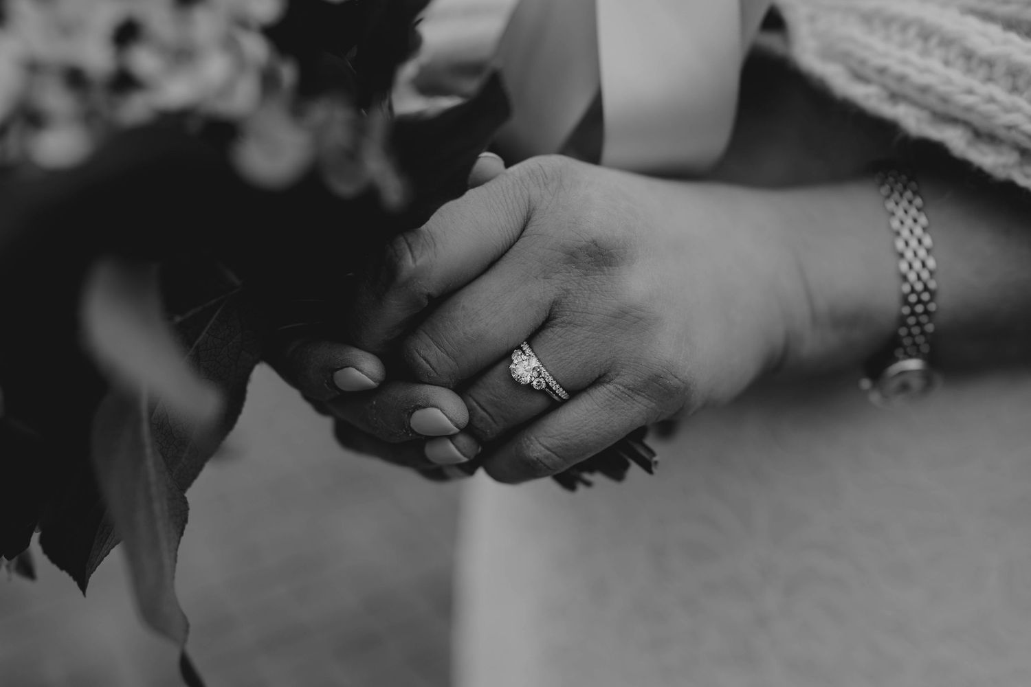 Black and white close-up of bride and groom holding hands, showing wedding ring and bracelet.