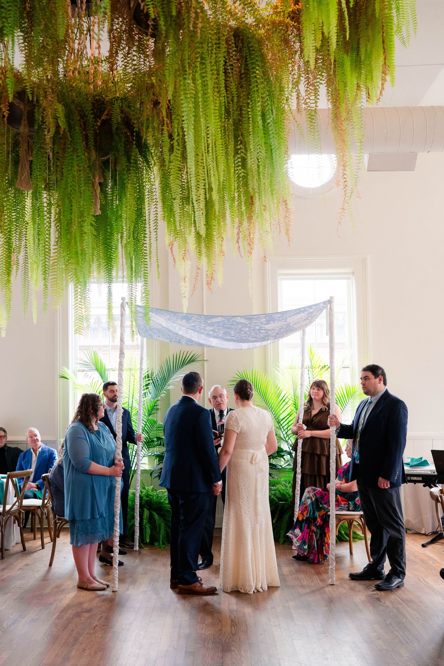 Intimate wedding ceremony beneath lush hanging ferns and blue chuppah in bright elegant venue.
