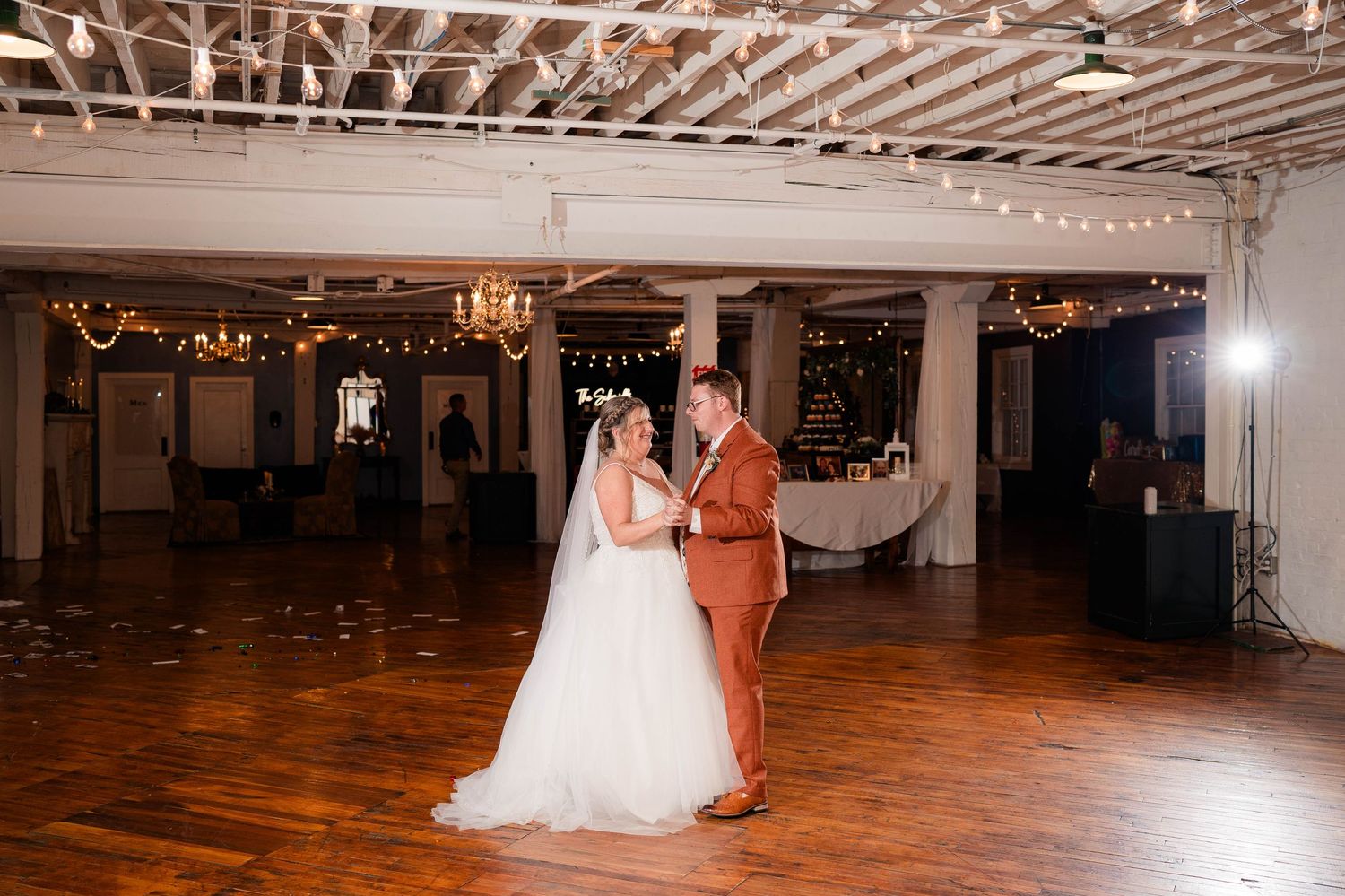 Bride and groom sharing first dance on wooden dance floor in warmly lit wedding reception venue.