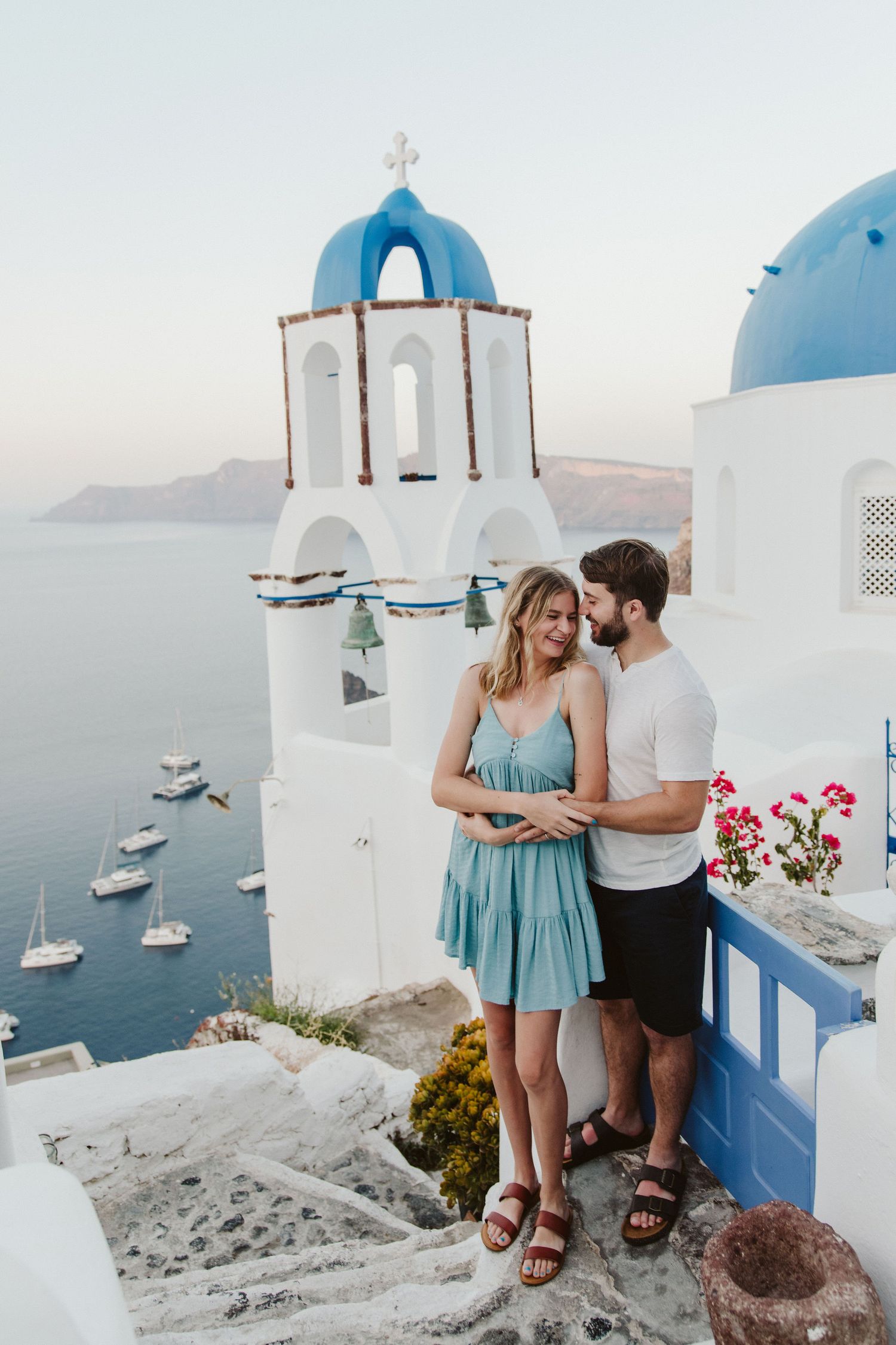 Couple embracing on steps in Santorini with iconic blue domed church and sea view behind them.