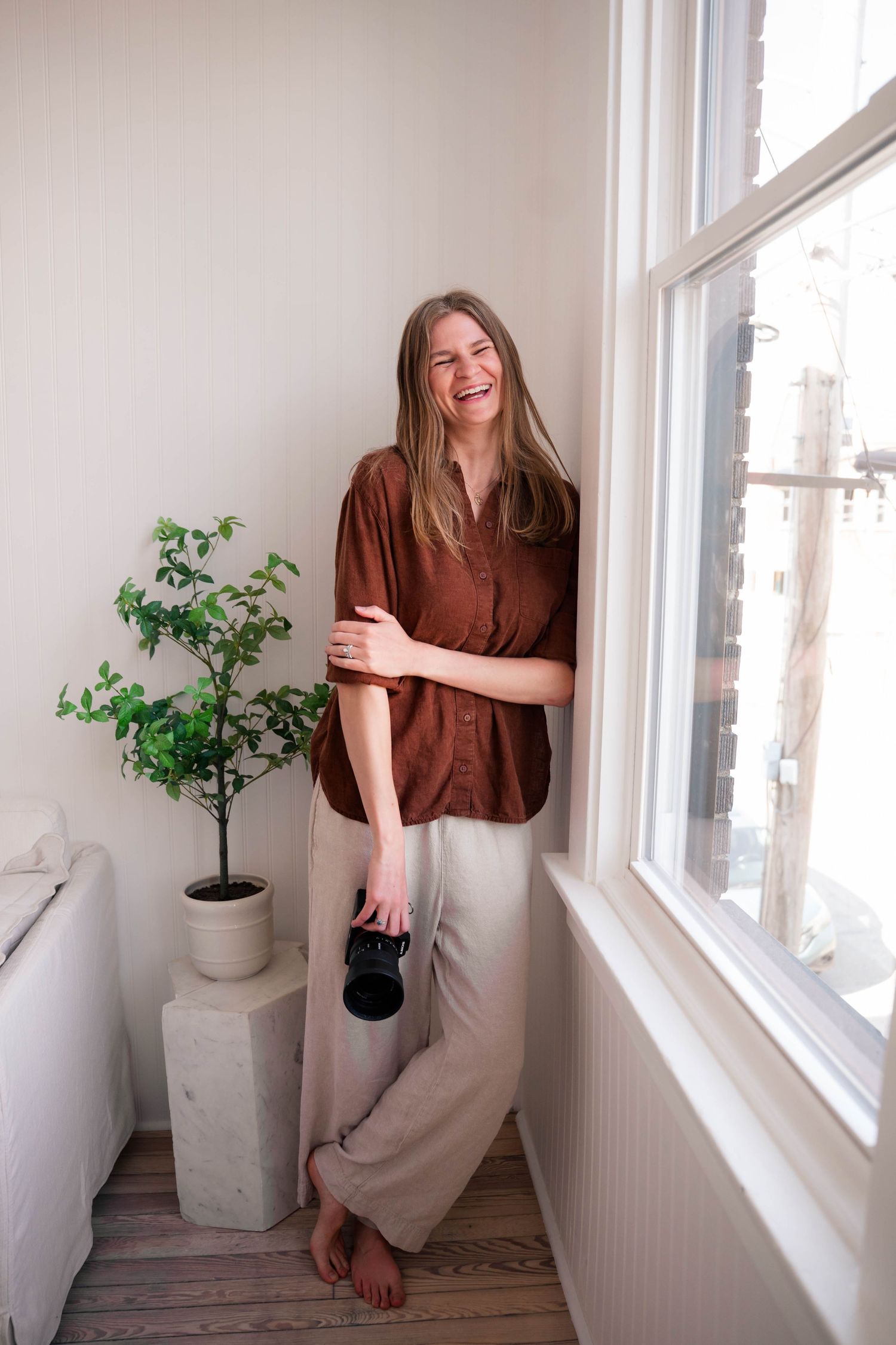Laughing woman in brown blouse holding camera standing barefoot by bright window with potted plant.