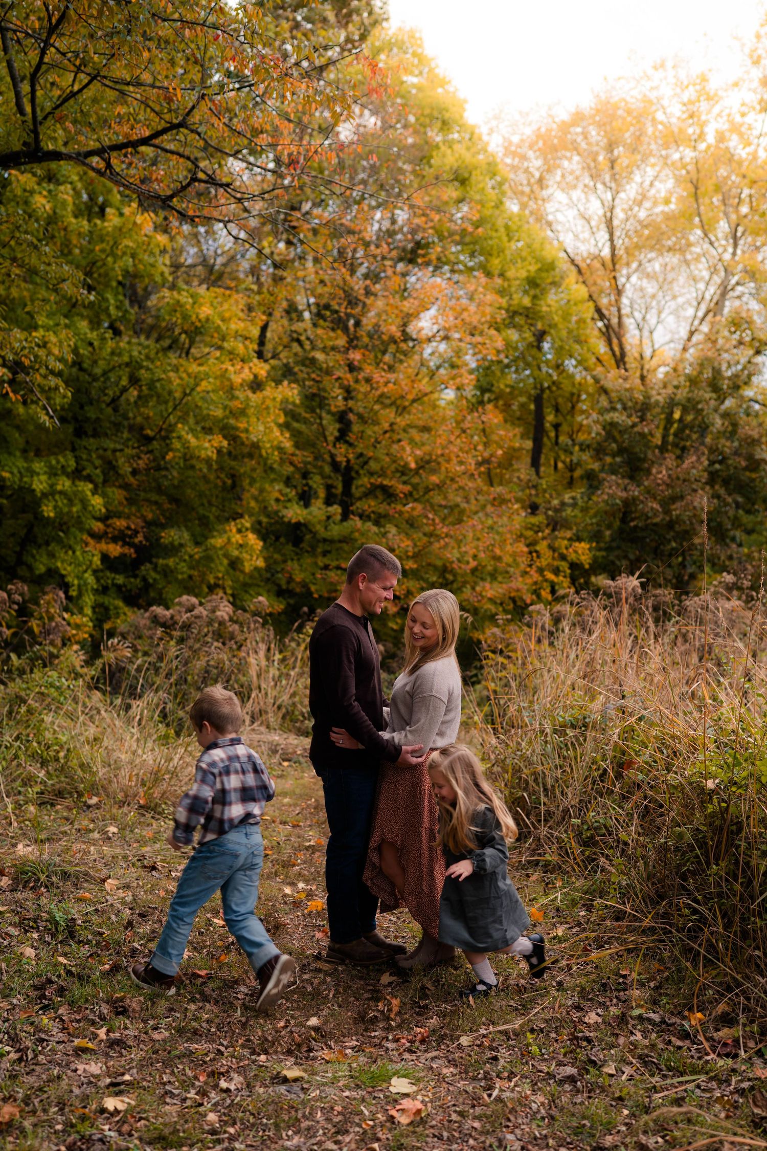 Family of four enjoying autumn walk in colorful fall forest, parents embracing while children play around them.
