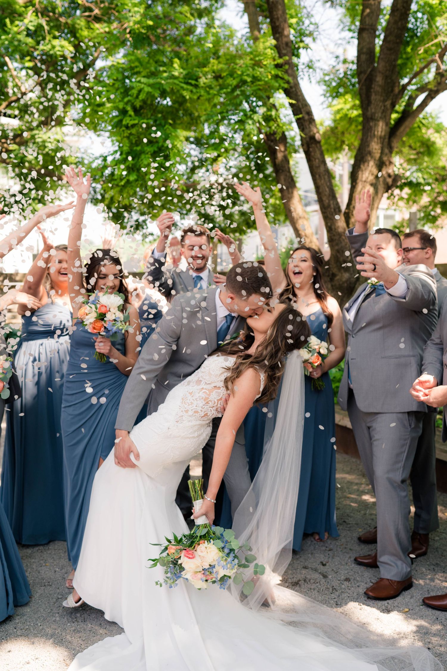 Wedding party celebrates with confetti as bride and groom share a romantic dip kiss outdoors.