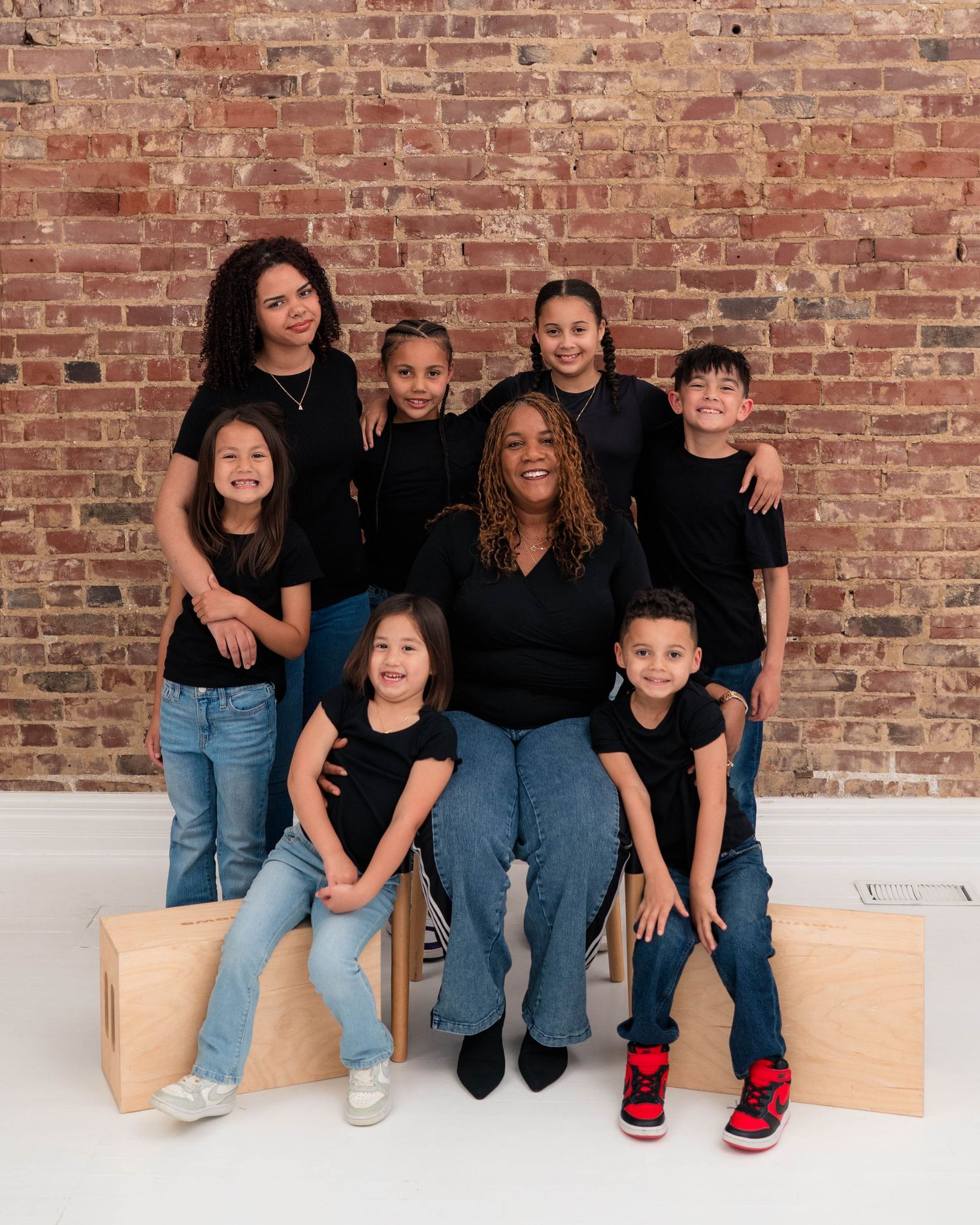 A joyful family portrait of a woman and six children in matching black tops and jeans posed in front of a brick wall.