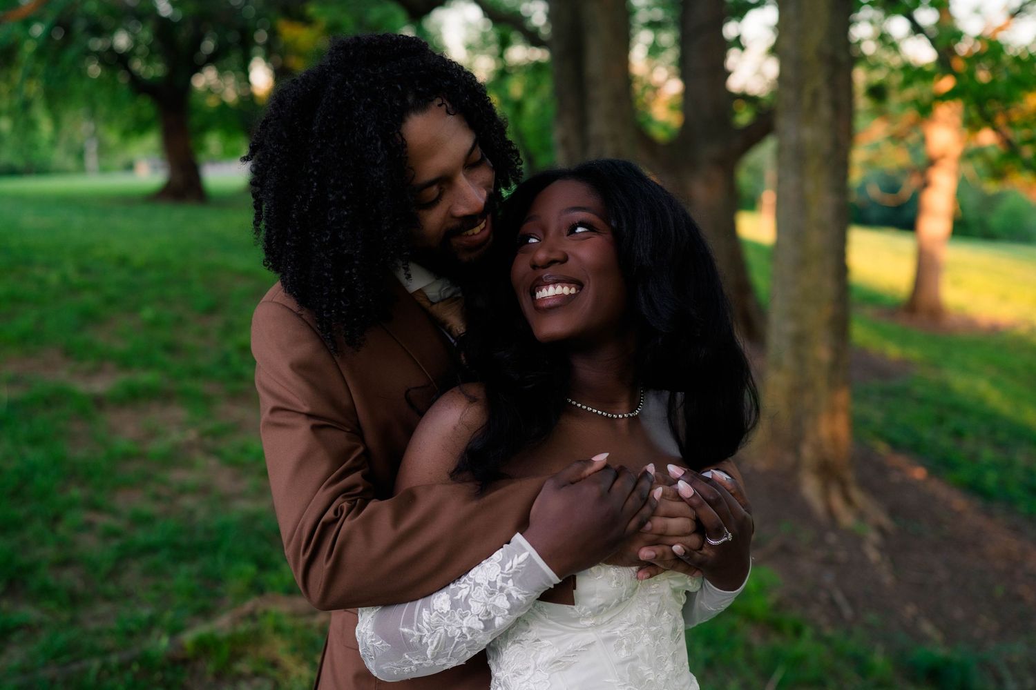 A joyful Black couple embraces outdoors, bride in white lace gown and groom in brown suit, surrounded by lush green trees.