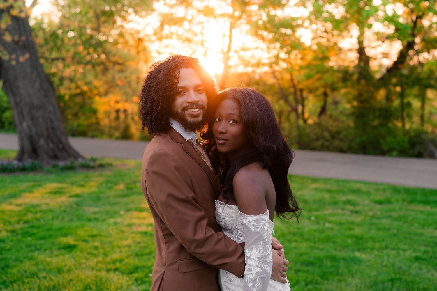 Smiling newlyweds embrace at golden sunset in lush green park, bride in lace gown.