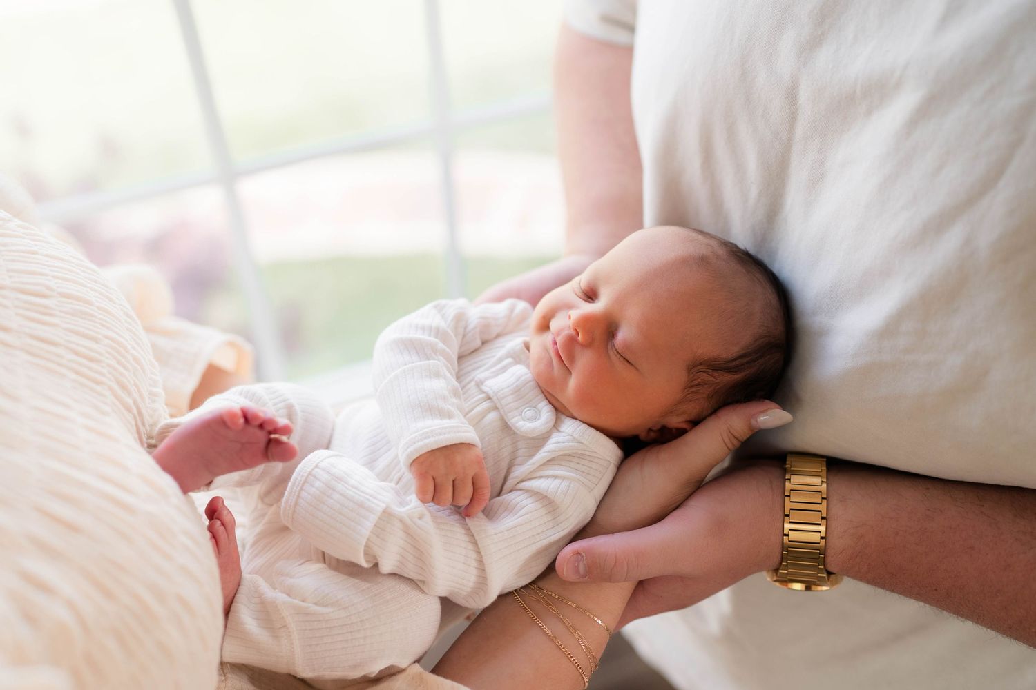 Newborn baby sleeping peacefully in parent's arms, wearing white onesie, with adult wearing gold watch visible.