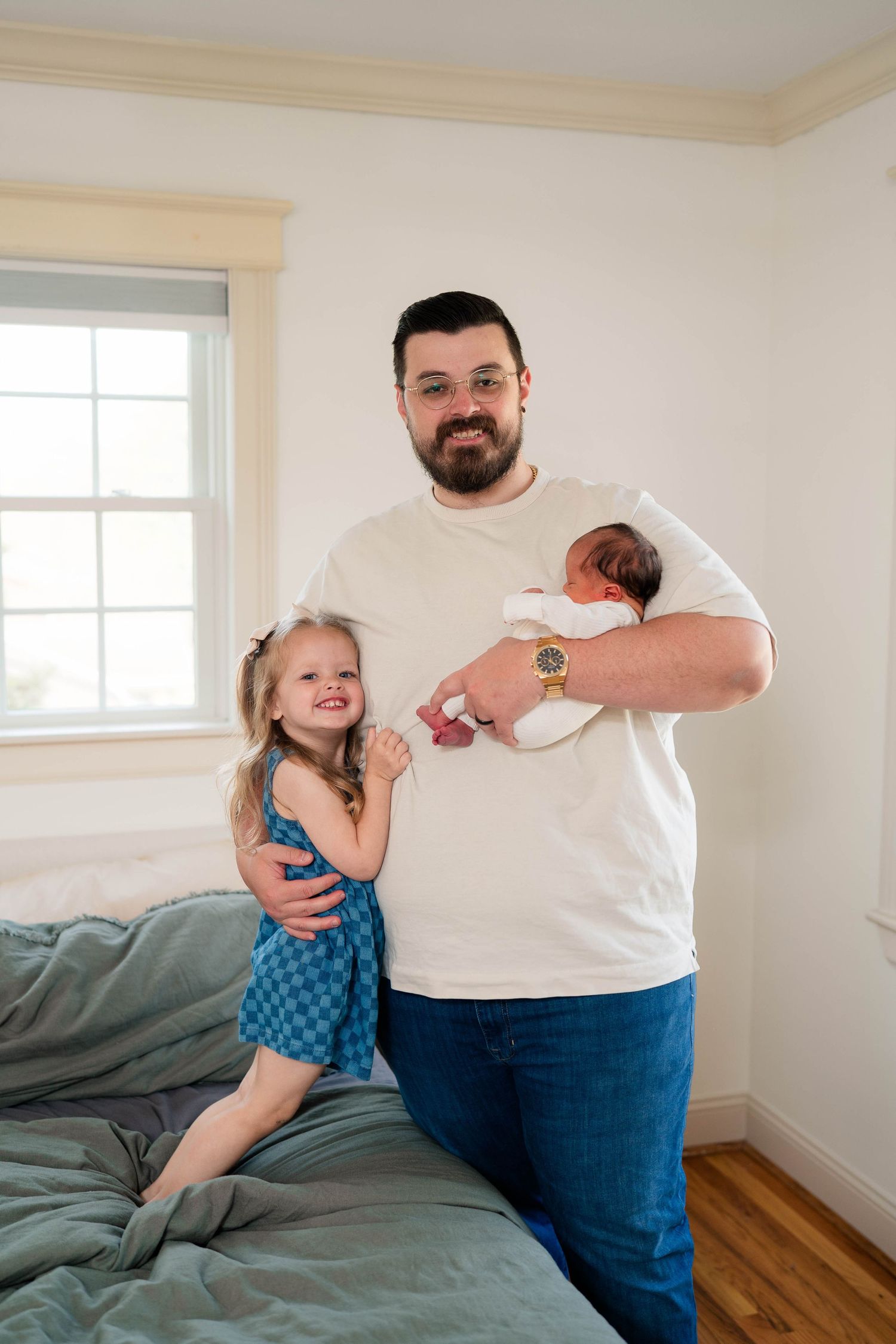 Proud father standing with toddler daughter and newborn baby in a bright bedroom setting.