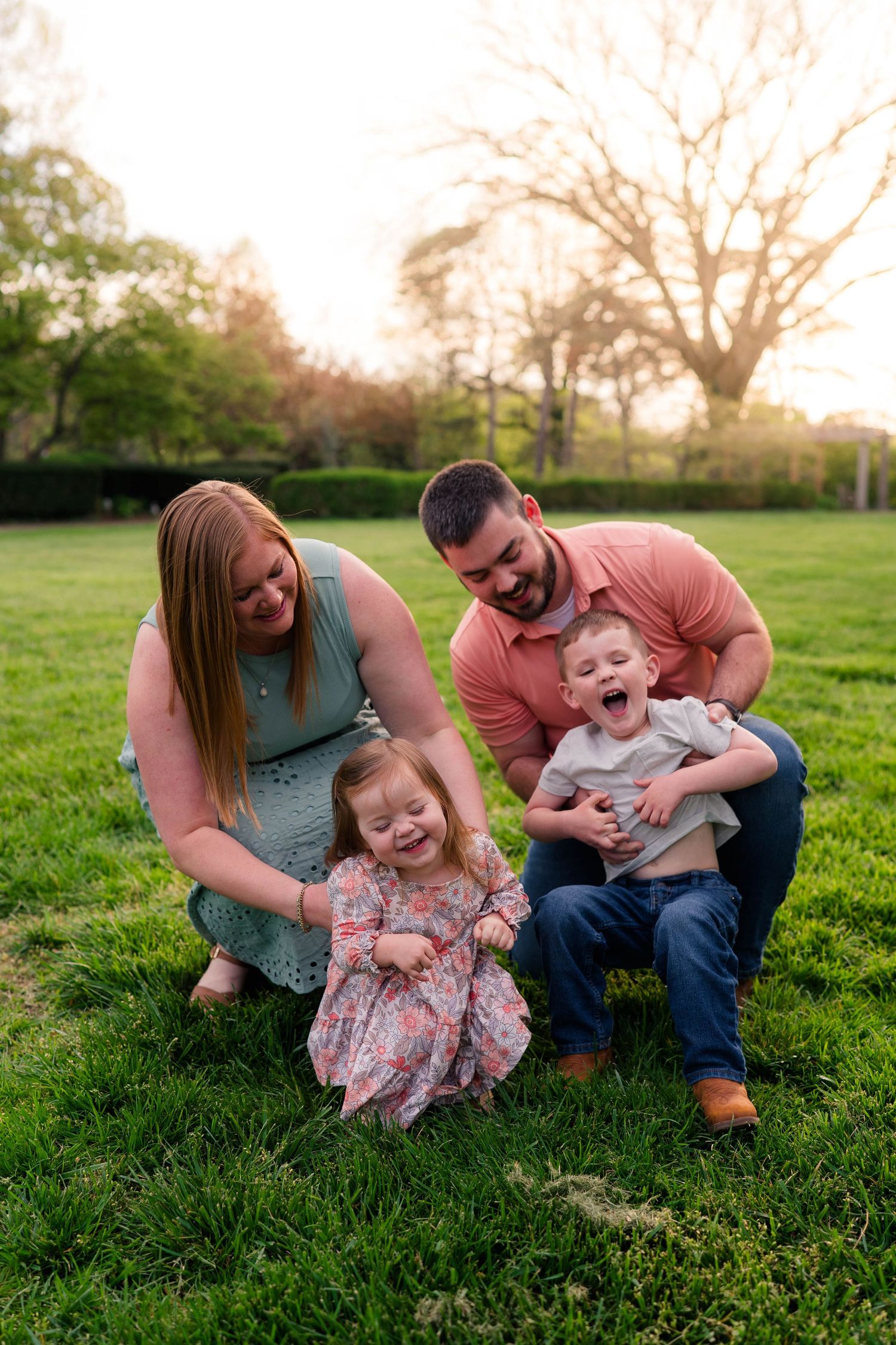 Happy family of four laughing together on green grass during golden hour in a park.