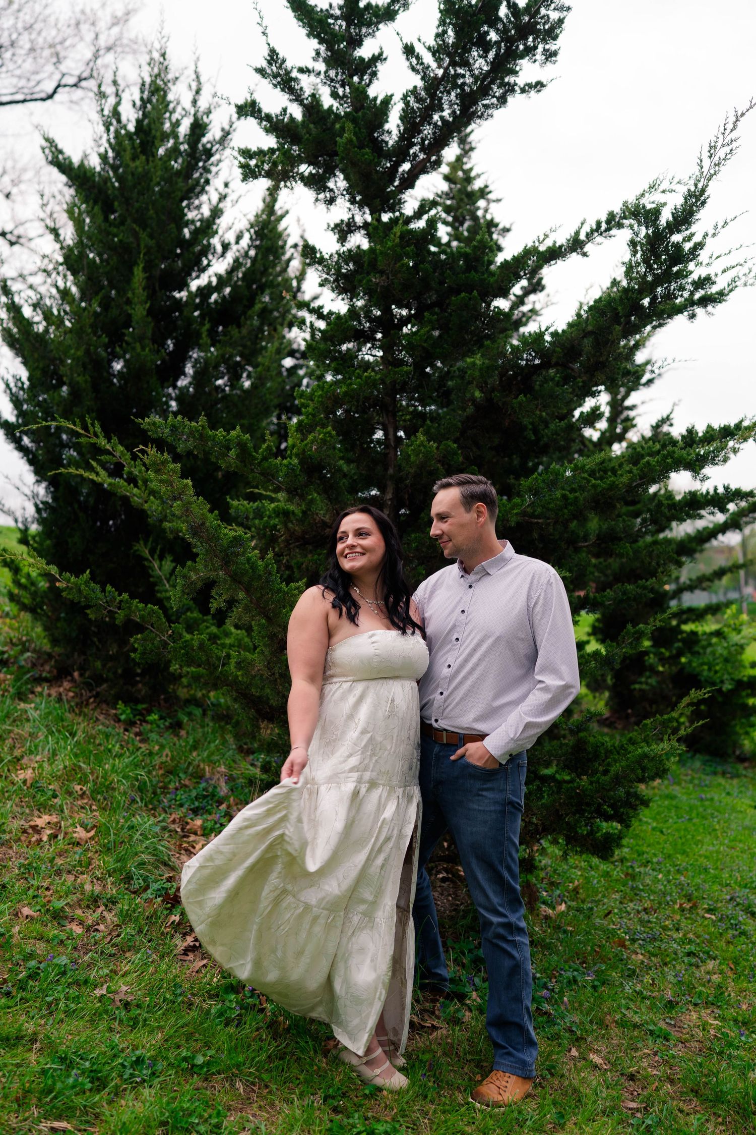 Couple standing together on grass in front of tall evergreen trees, woman in flowing white dress.