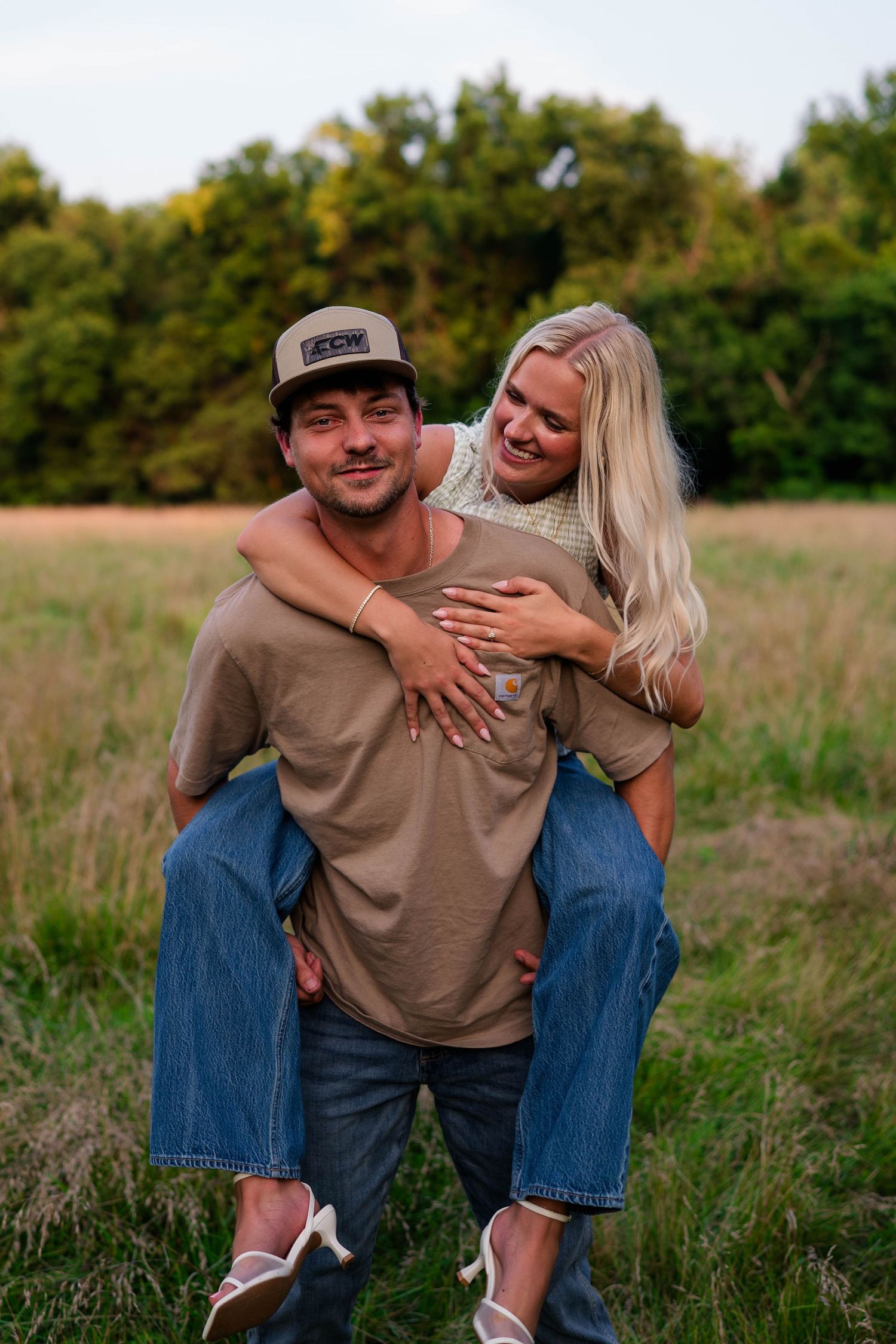 A smiling man gives his girlfriend a piggyback ride in an open field, both laughing and wearing casual clothes.