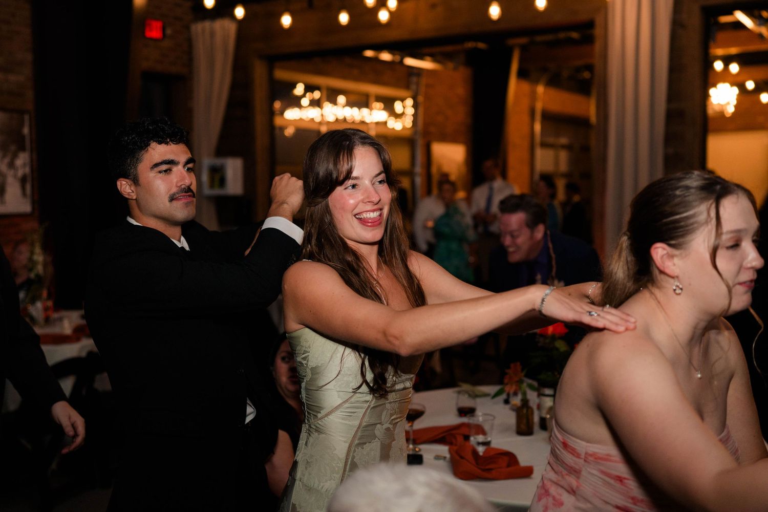 Laughing wedding guest reaches across table in animated conversation during lively rustic wedding reception dinner.