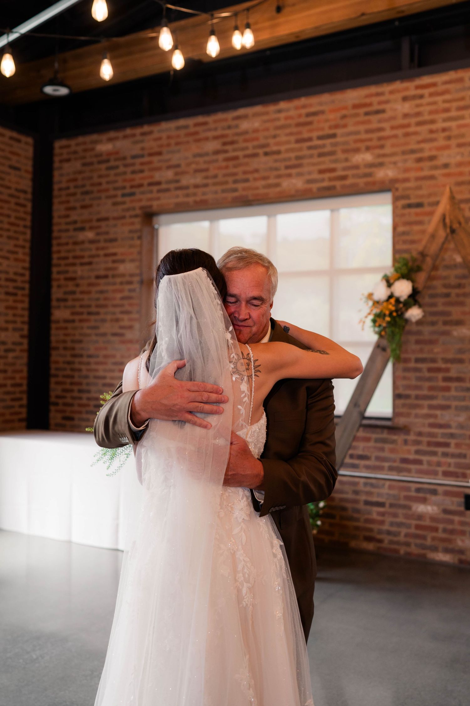 Father and bride share an emotional hug during a wedding reception in a rustic brick-walled venue.