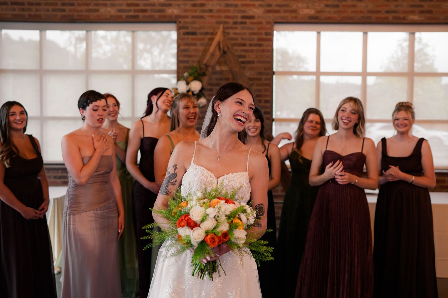 Laughing bride in white gown holds colorful bouquet as bridesmaids in mismatched dresses react joyfully behind her.
