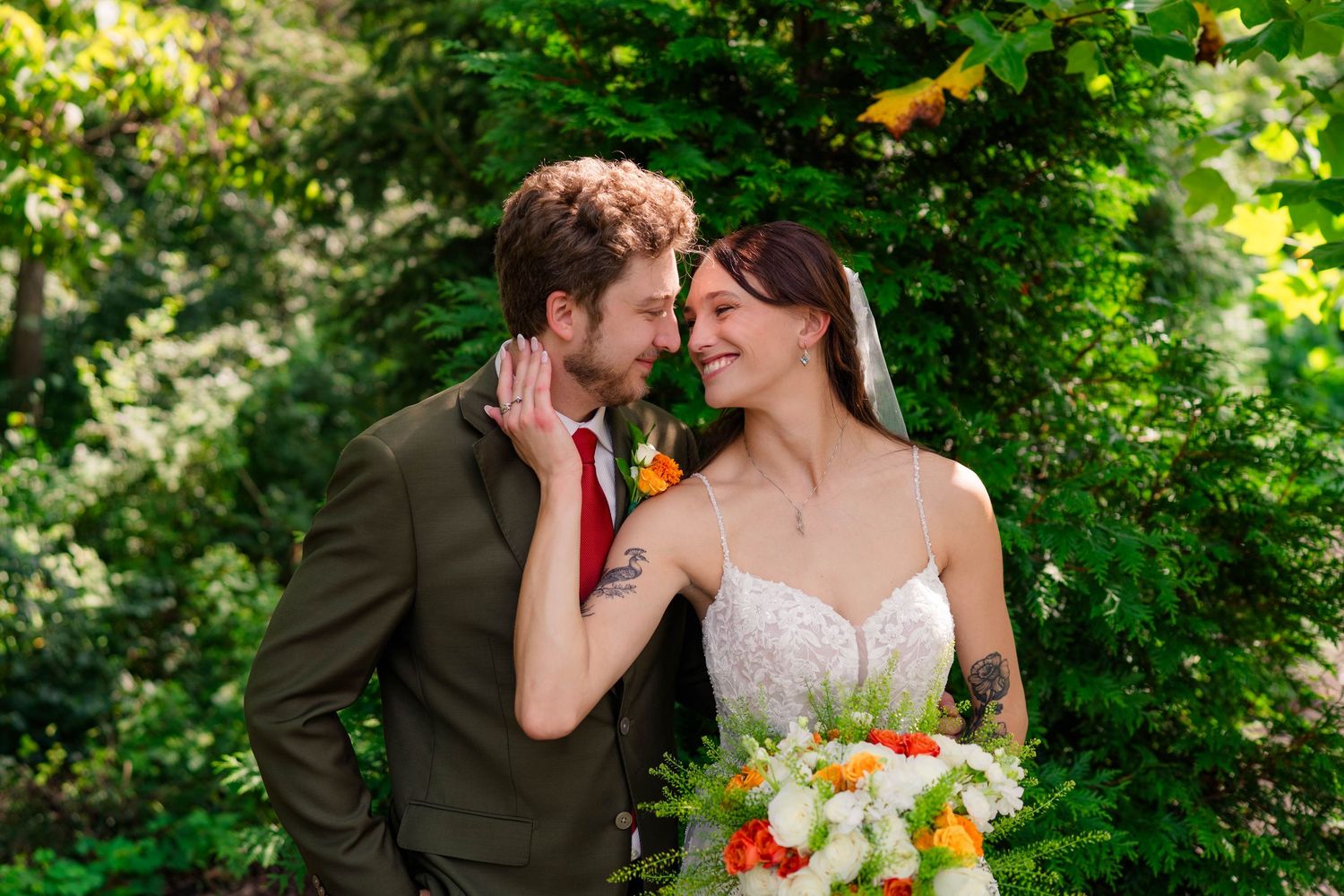 Bride and groom share an intimate moment surrounded by lush green foliage, bride holding colorful bouquet.