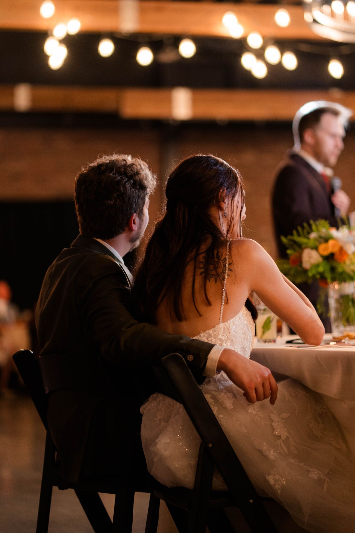 Bride and groom sit together at sweetheart table under string lights during intimate wedding reception dinner.