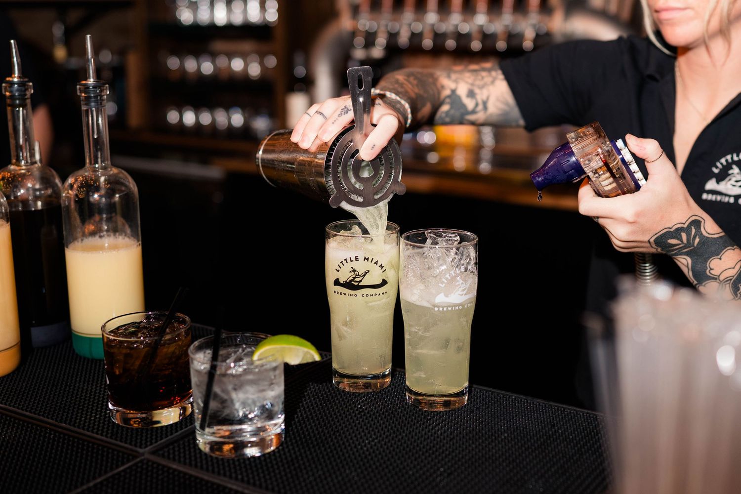 Tattooed bartender pours craft cocktails into glasses at stylish wedding reception bar.