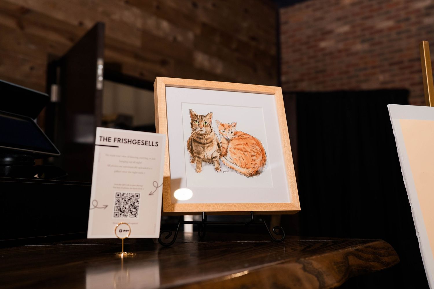Framed watercolor painting of two cats displayed on easel at wedding reception with QR code sign.