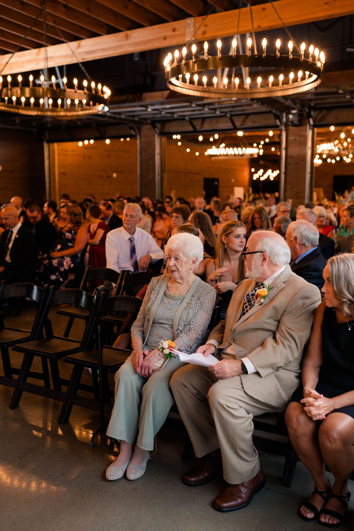 Wedding guests seated in chairs under chandelier lighting at indoor ceremony venue.