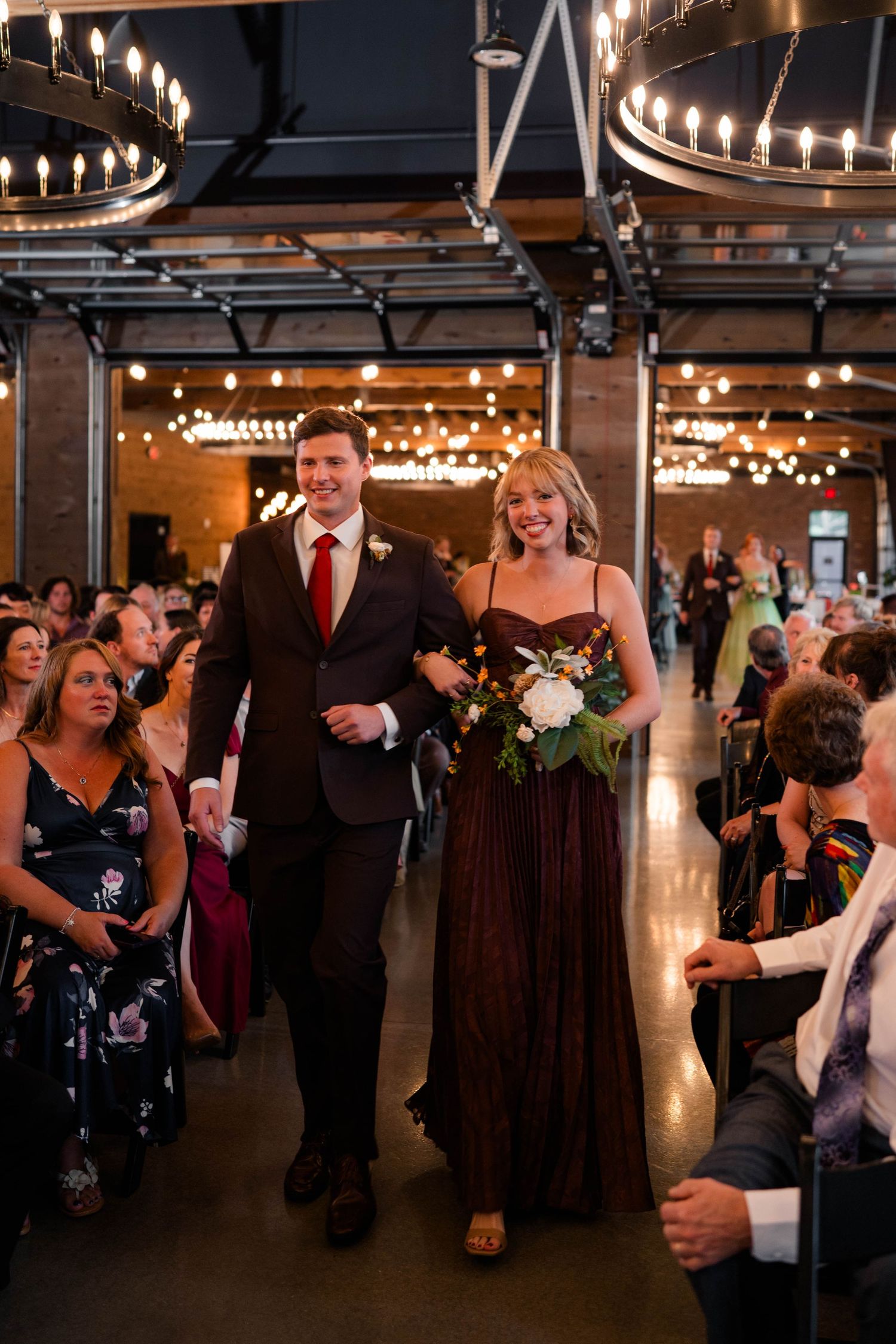Bridesmaid in burgundy gown walks down aisle with groomsman at elegant indoor wedding.