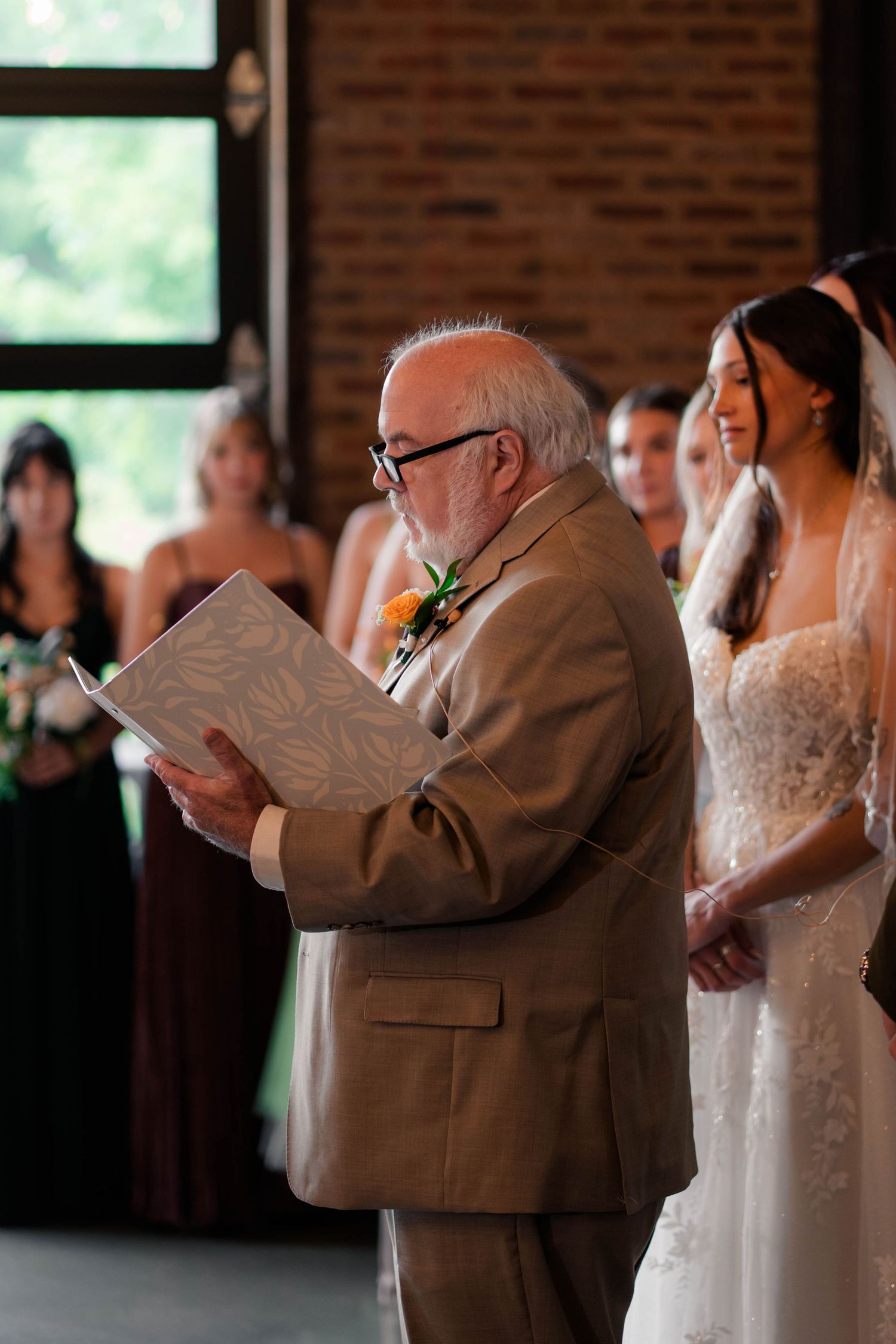 Elderly officiant reads from a book during indoor wedding ceremony as bride smiles beside him.