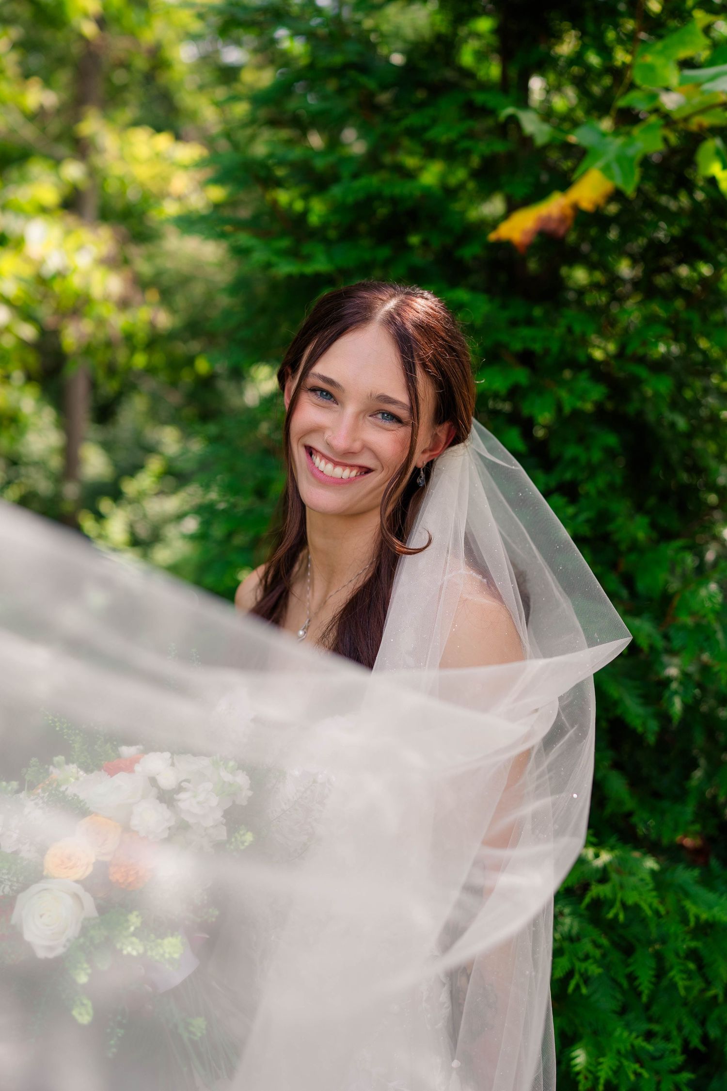 Radiant bride smiling behind flowing veil surrounded by lush green trees on wedding day.