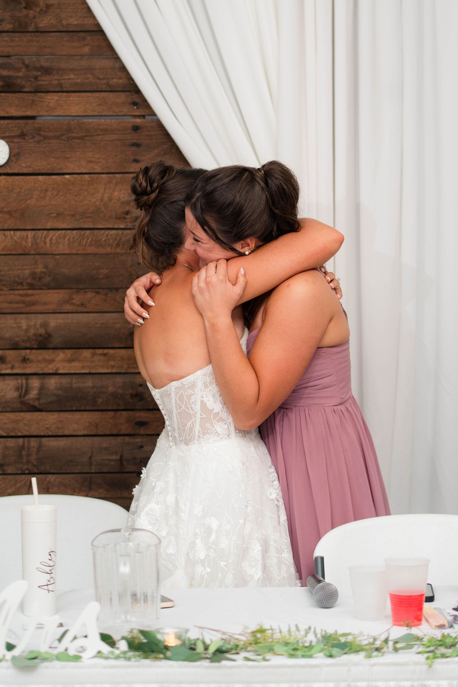 Bride and bridesmaid share an emotional hug at wedding reception near rustic wood wall backdrop.