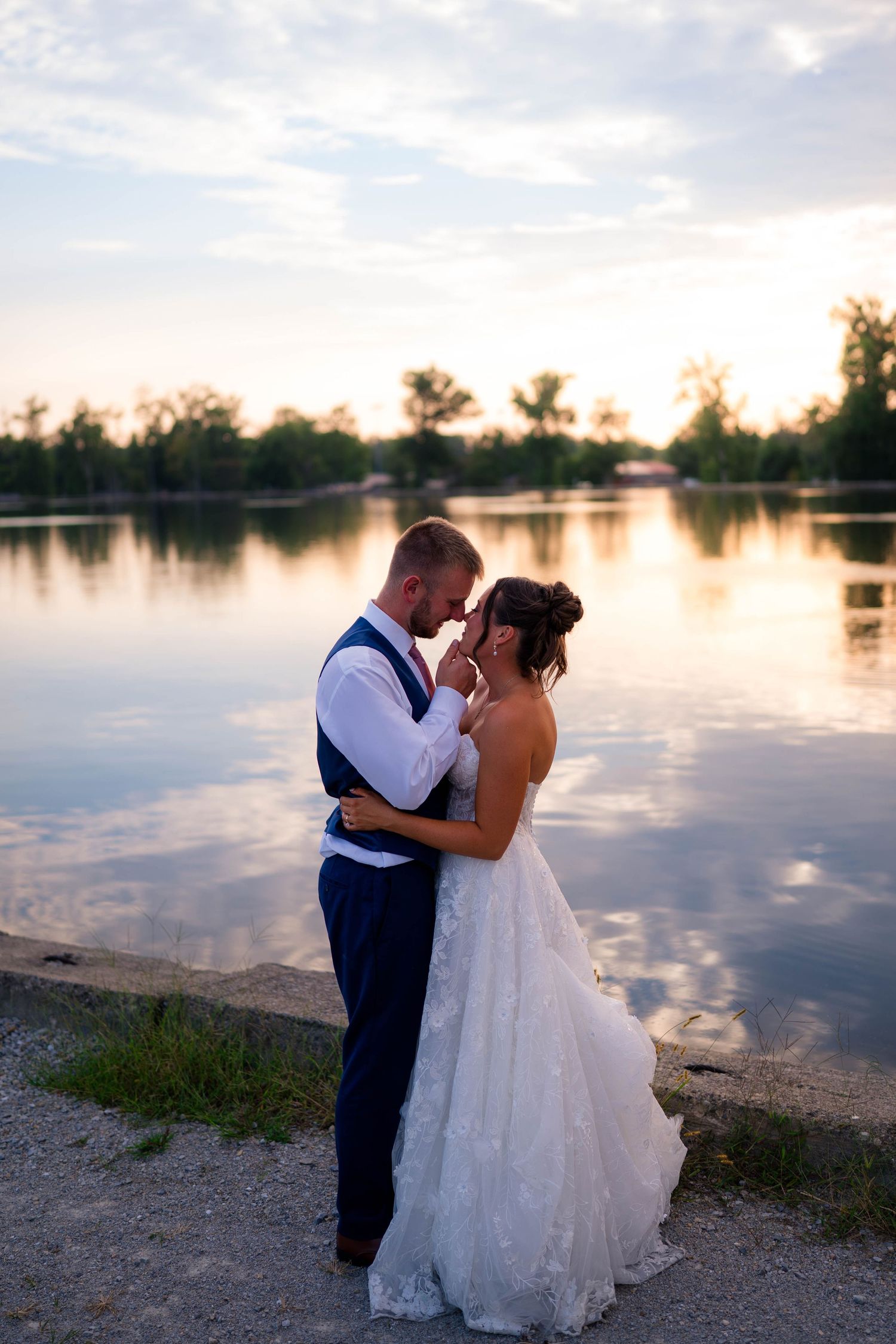 Bride and groom share a romantic forehead touch by a reflective lake at sunset.