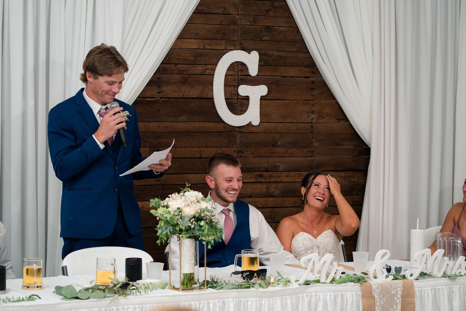 A groomsman delivers a humorous wedding speech as the bride laughs and groom smiles at a rustic reception venue.
