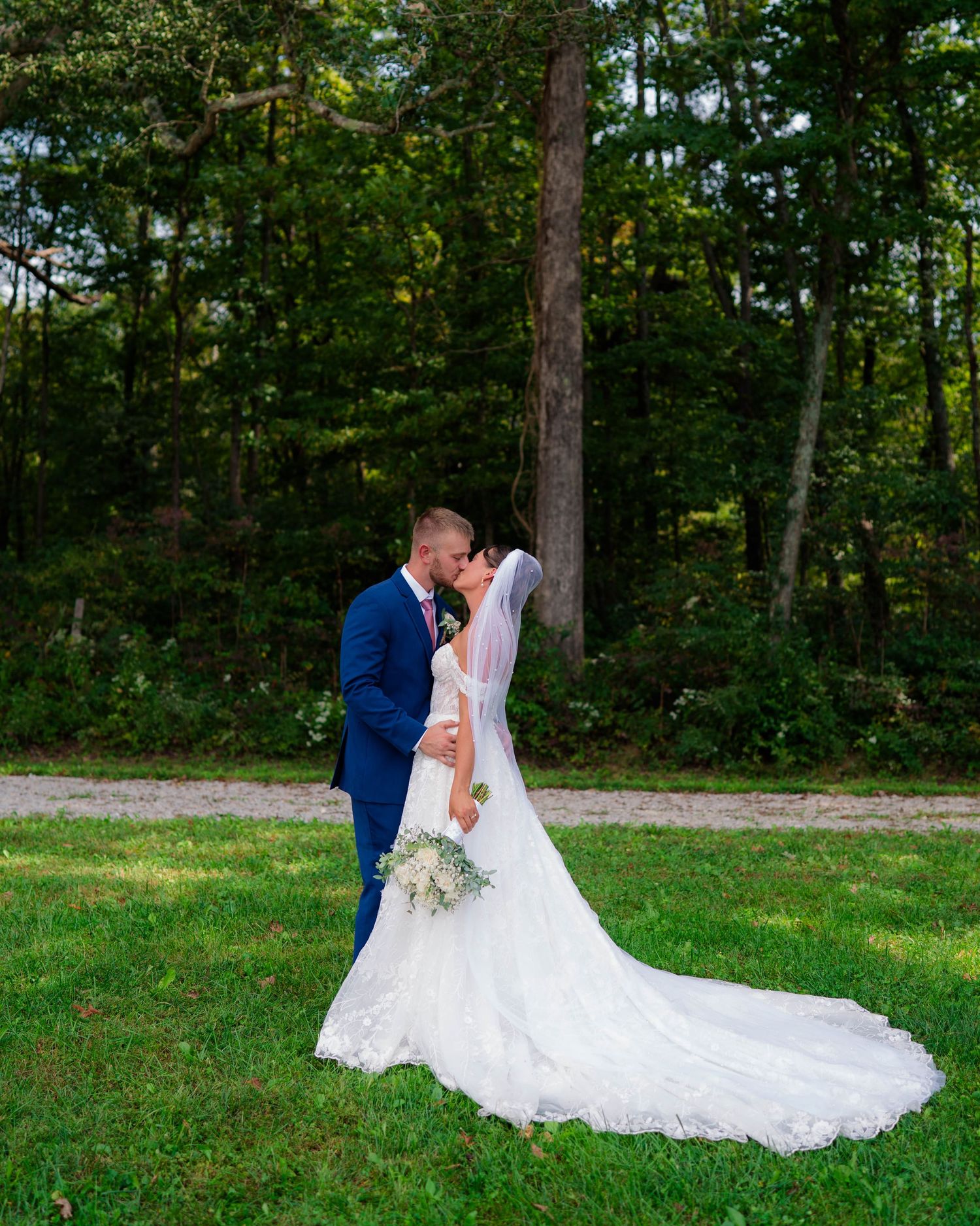 Bride and groom sharing a romantic kiss on green grass with tall trees in the background.