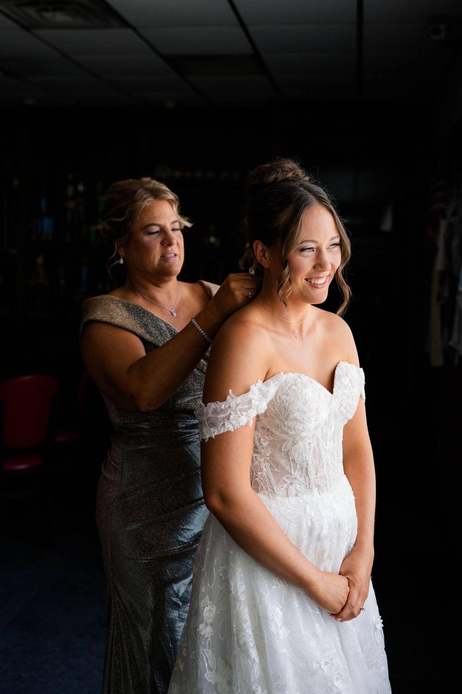 Mother helping smiling bride with earrings as she prepares in her lace off-shoulder wedding gown.
