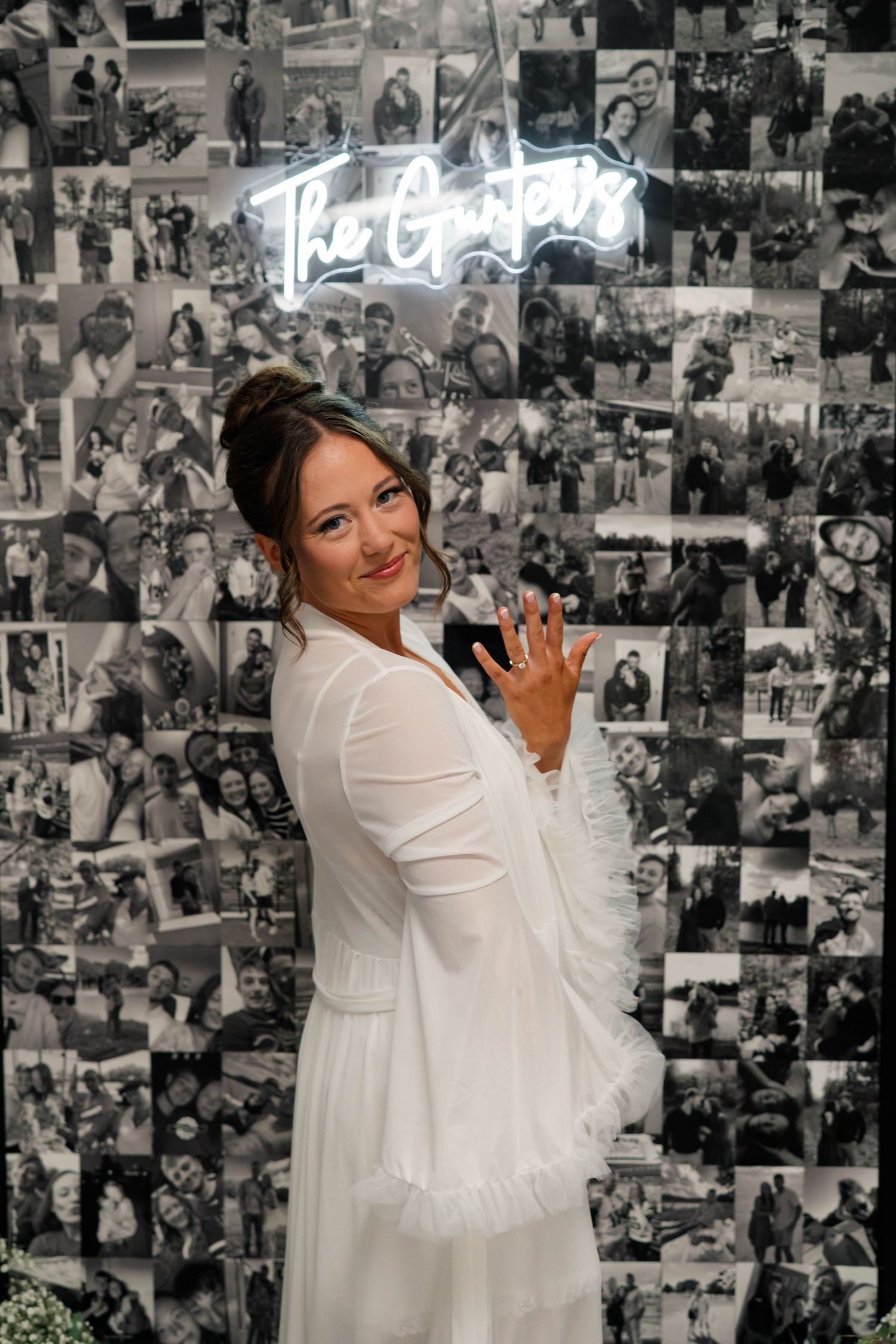 Bride in white dress posing before black and white photo collage wall with neon sign.