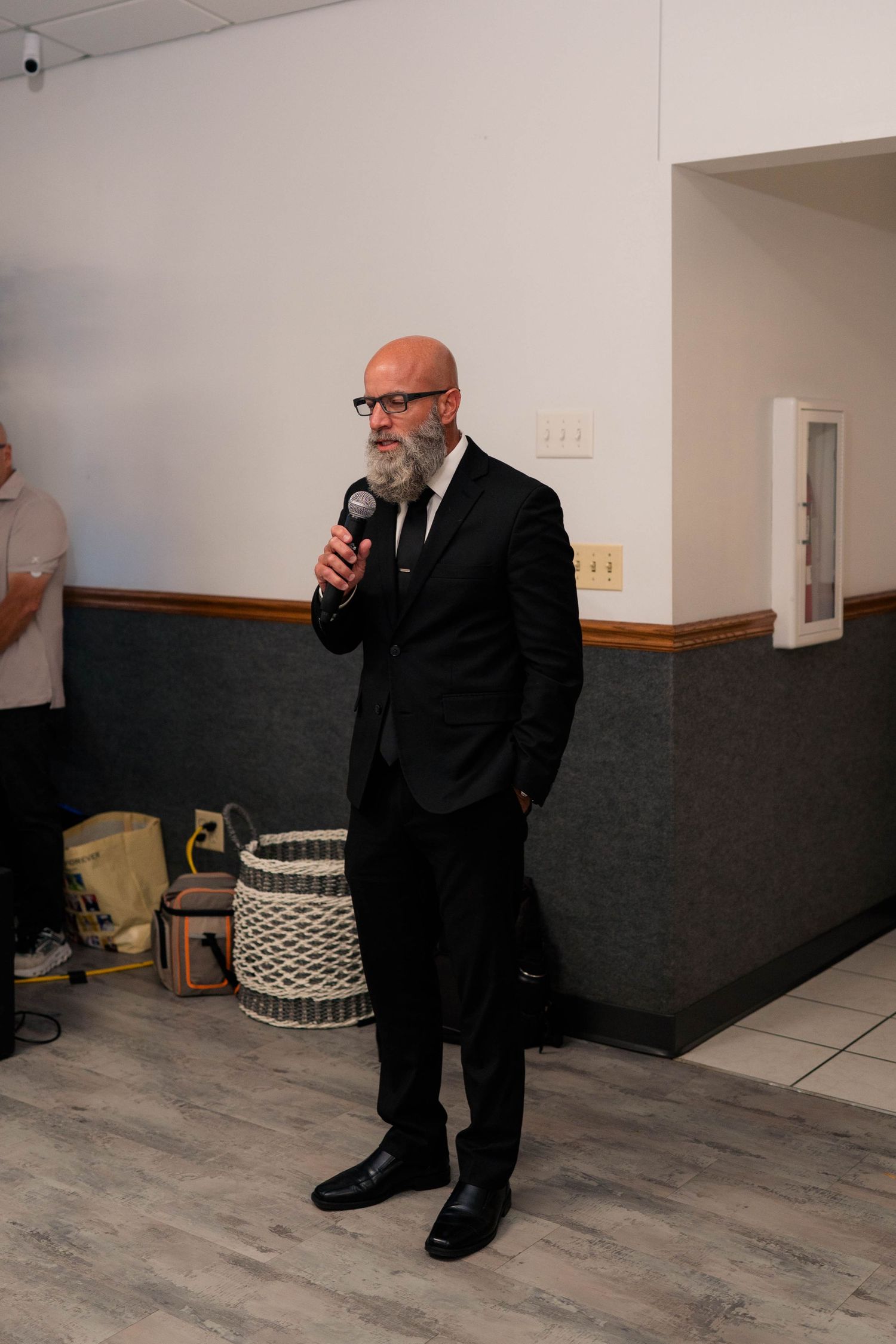 Bald man with gray beard in black suit speaks into microphone at indoor wedding reception event.