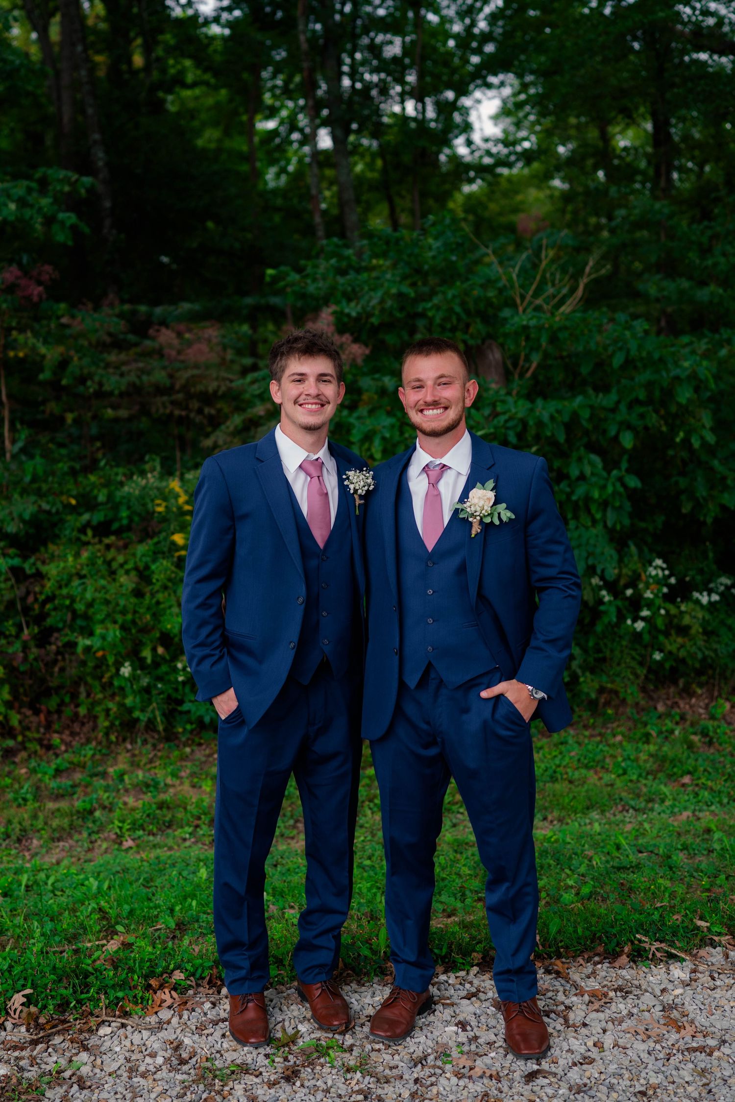 Two groomsmen in navy blue suits with pink ties and boutonnieres smiling outdoors at wedding.