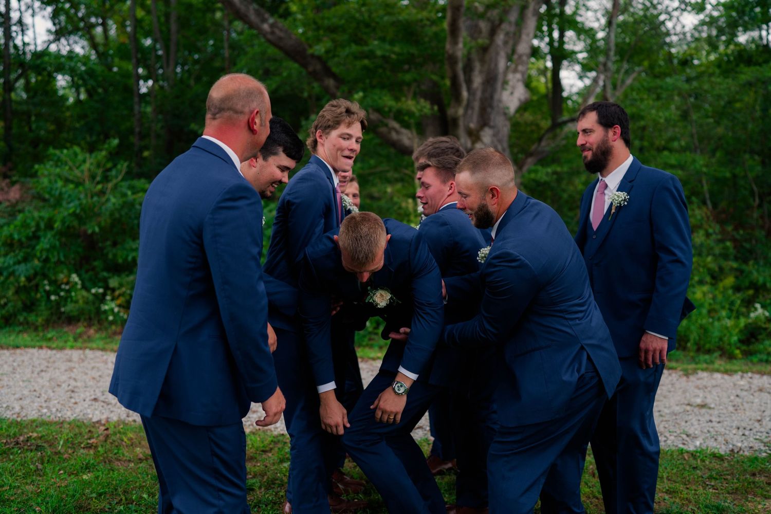 Groomsmen in navy suits playfully roughhouse together outdoors in a wooded area during wedding celebrations.
