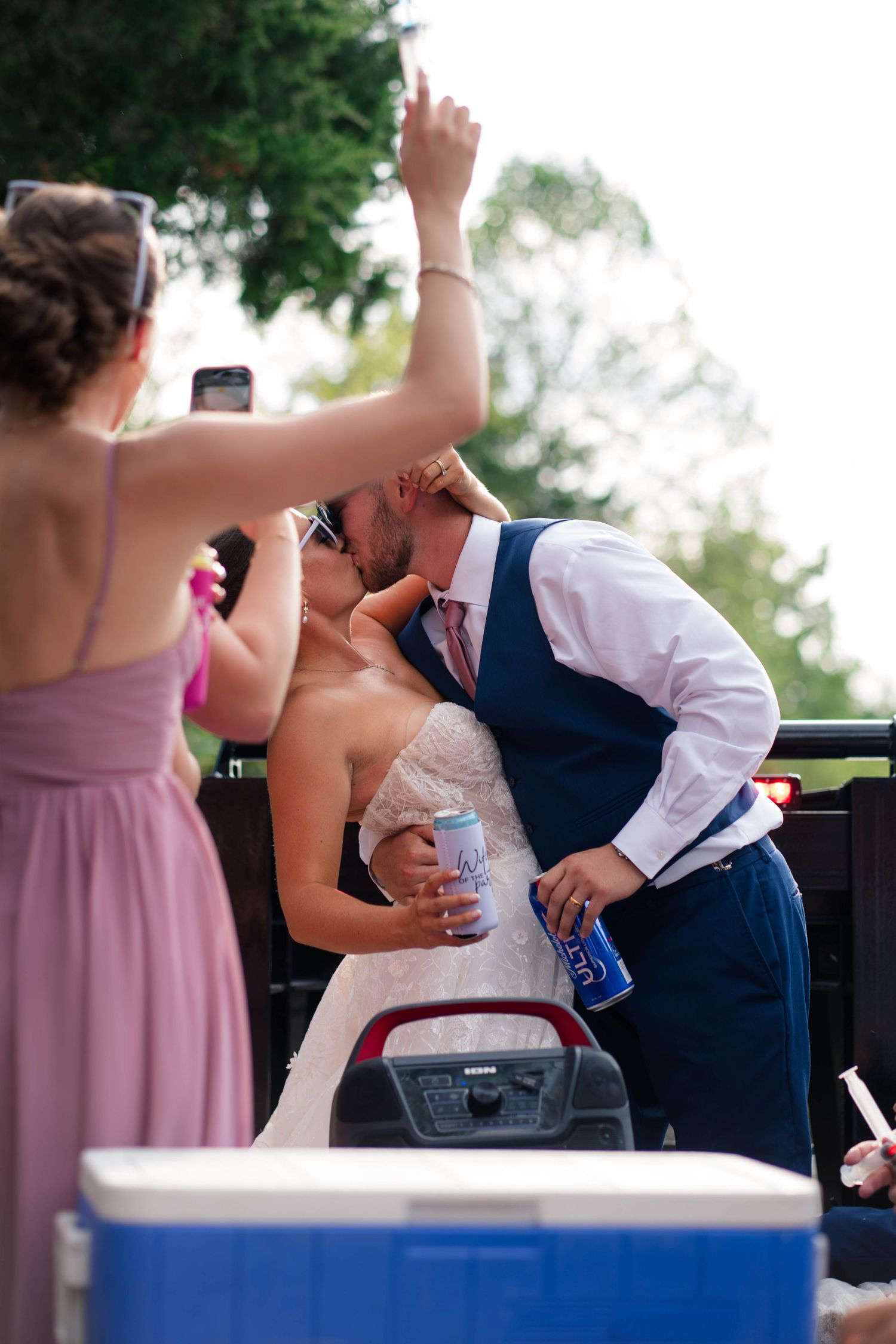Bride and groom share a kiss on truck bed while bridesmaid in purple dress photographs the moment.