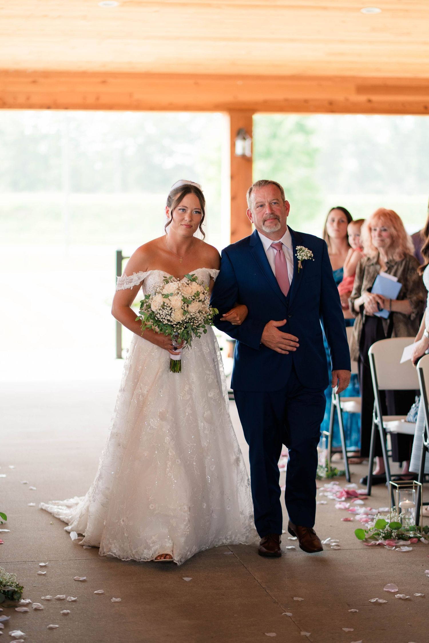 Bride in off-shoulder lace gown holding white bouquet walks down aisle with escort in navy suit.