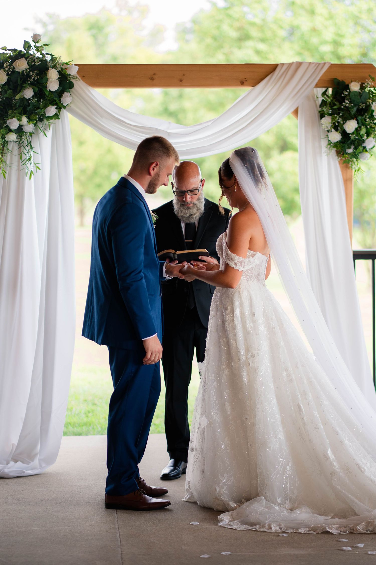 Bride and groom exchange rings with officiant under white draped wedding arch.