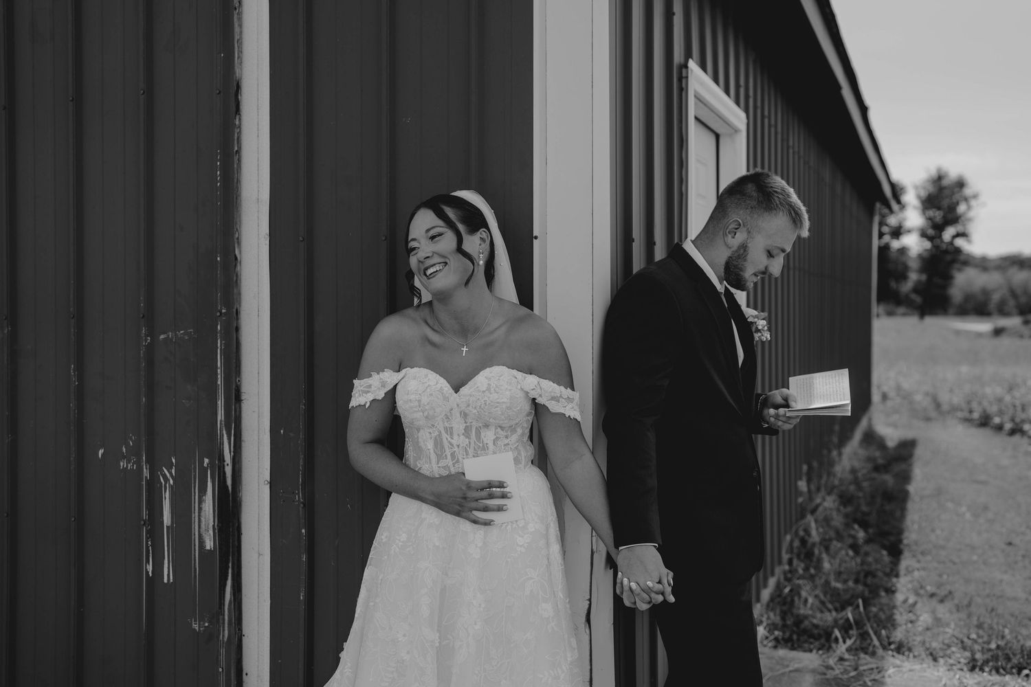 Black and white photo of bride and groom holding hands and smiling beside a red barn.