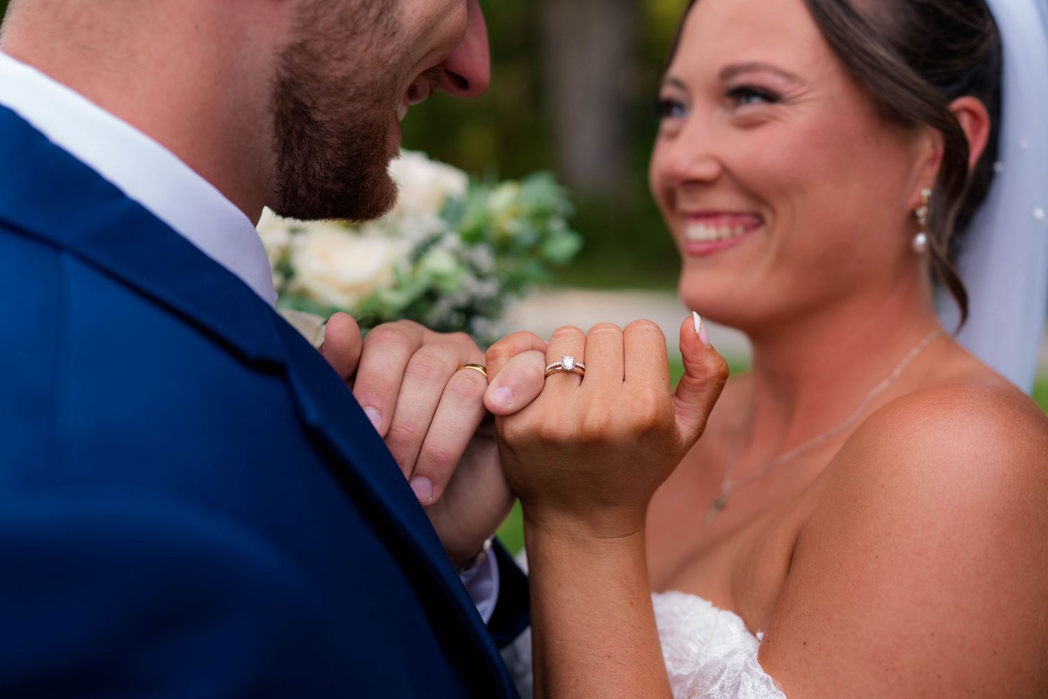 Smiling bride shows off her engagement ring while holding hands with her groom outdoors.