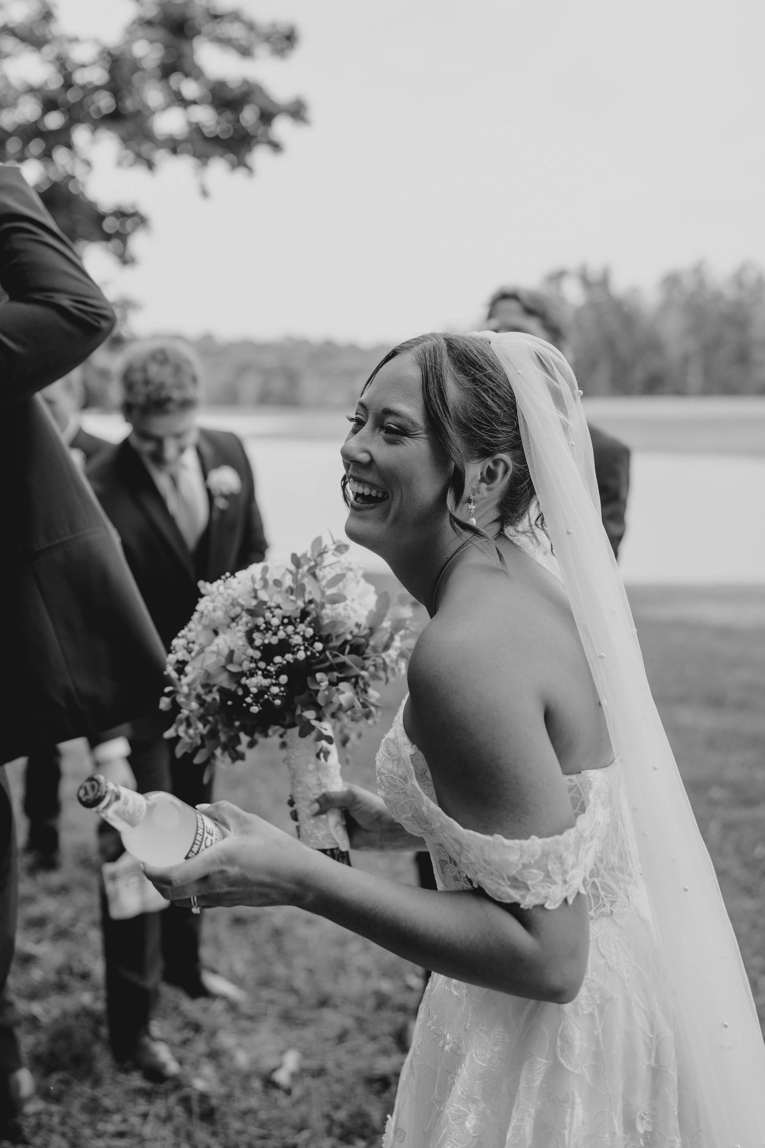 Laughing bride in off-shoulder lace gown holds bouquet and champagne bottle in black and white wedding photo.