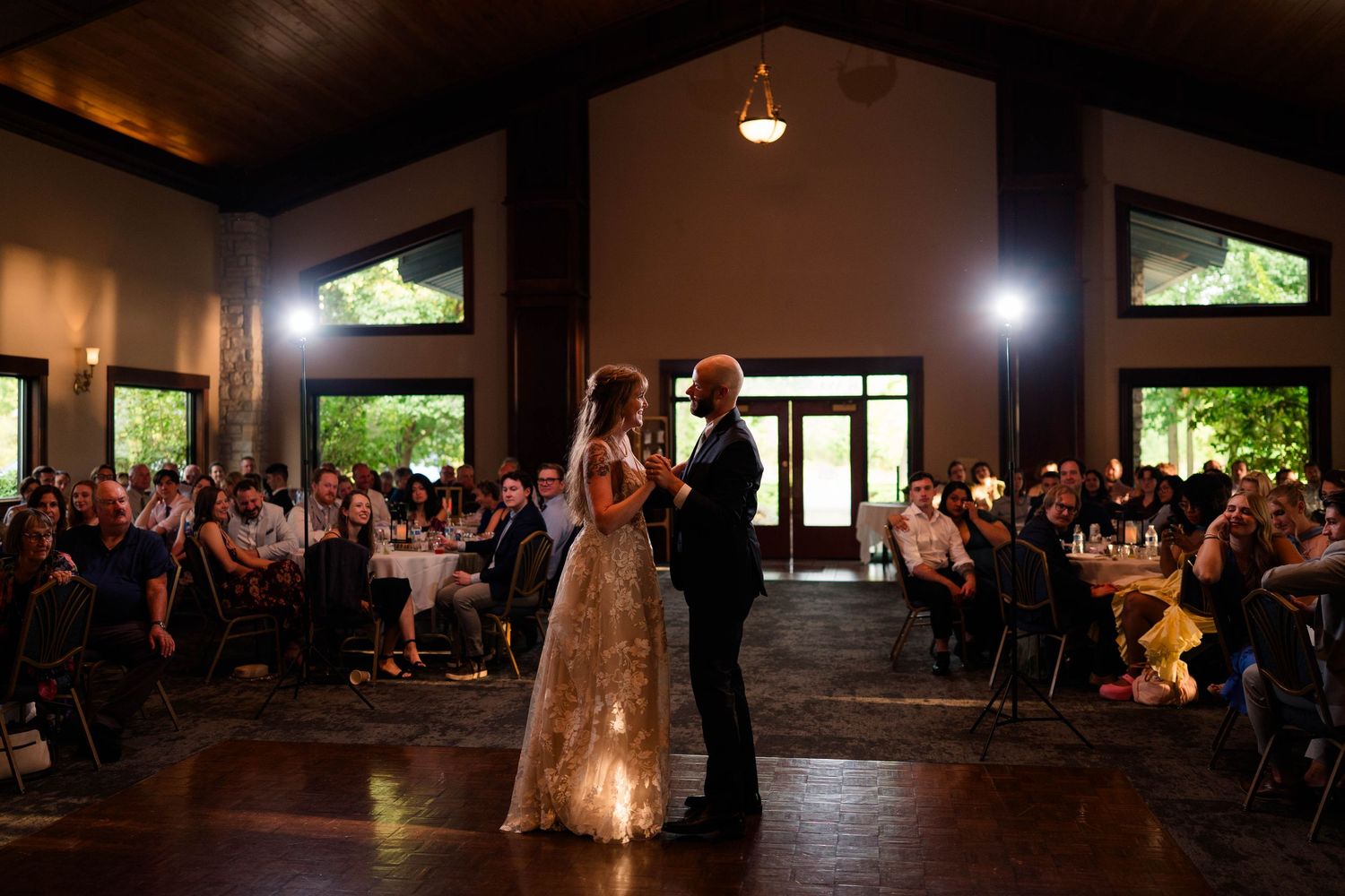 Bride and groom share first dance before seated wedding guests in a rustic venue.